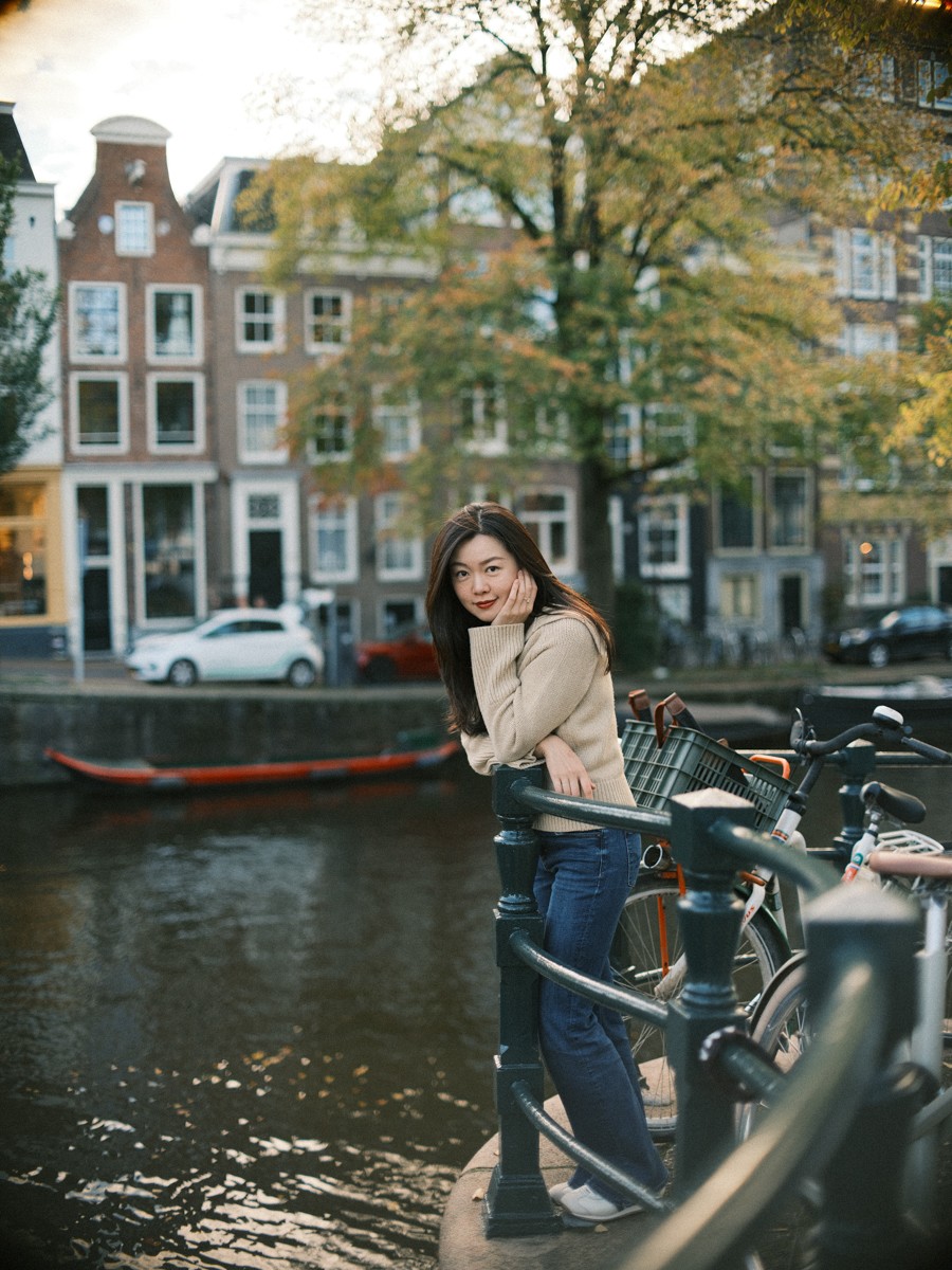 Vrouw rust met haar kin op haar hand bij een hek langs de gracht, met grachtenpanden en een vergelende boom in Amsterdam