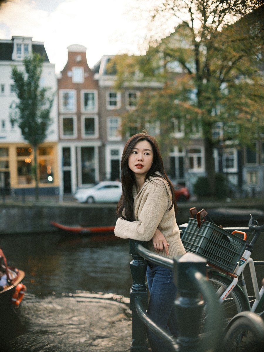 Soloreiziger leunt op een hek langs de gracht naast een fiets met een krat en kijkt om, met een rode boot op het water