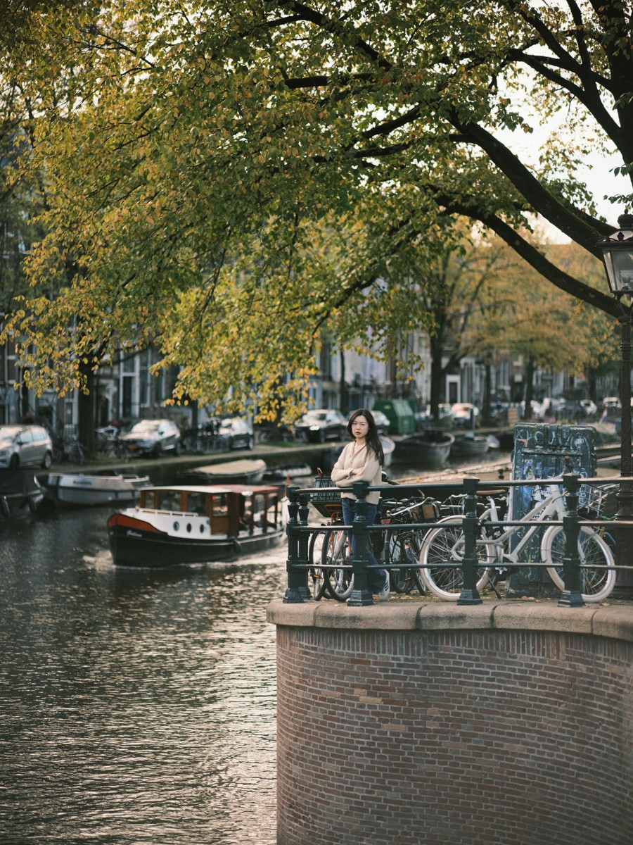 Kleine figuur van een vrouw op een hoek aan de gracht tussen geparkeerde fietsen, een grote boom boven het water en een varende boot