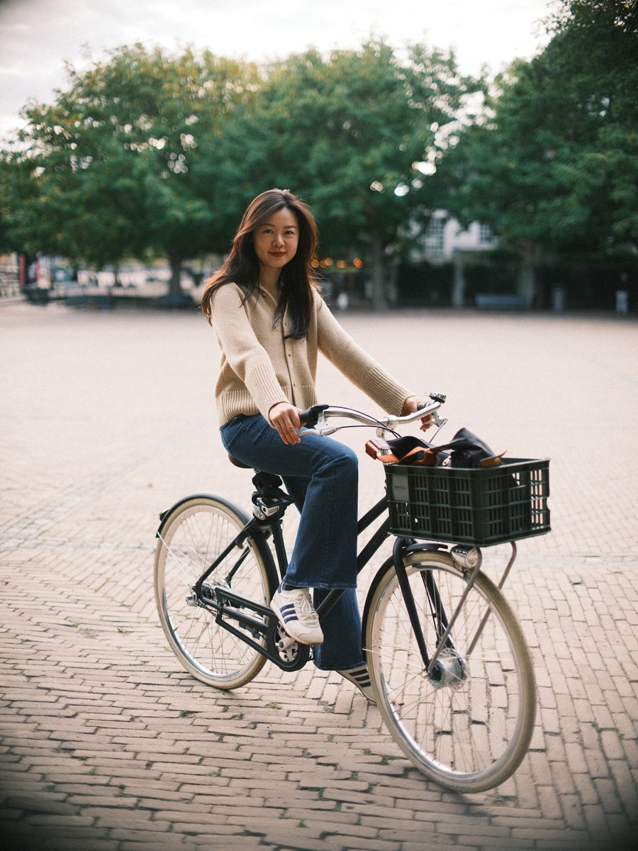 Soloreiziger fietst op een zwarte Hollandse fiets over een plein van klinkers in Amsterdam, lachend, haar tas in de krat voorop
