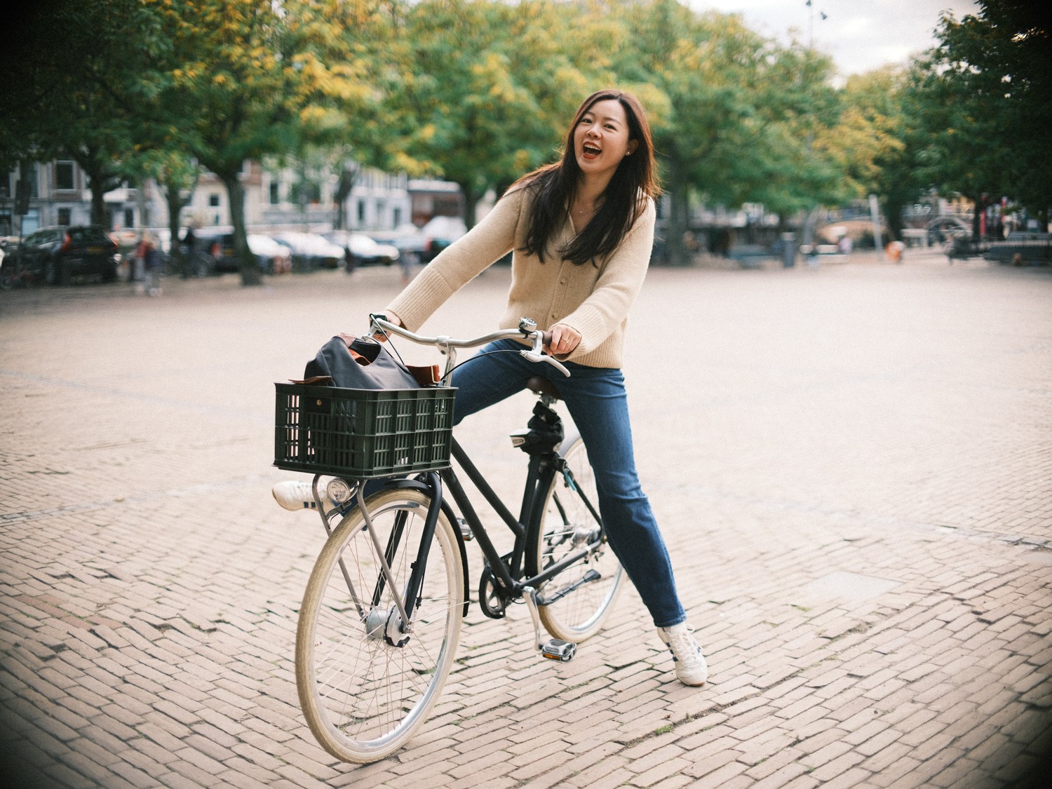 Soloreiziger lacht op een zwarte Hollandse fiets met een krat voorop, met een breed plein van klinkers en bomen erachter