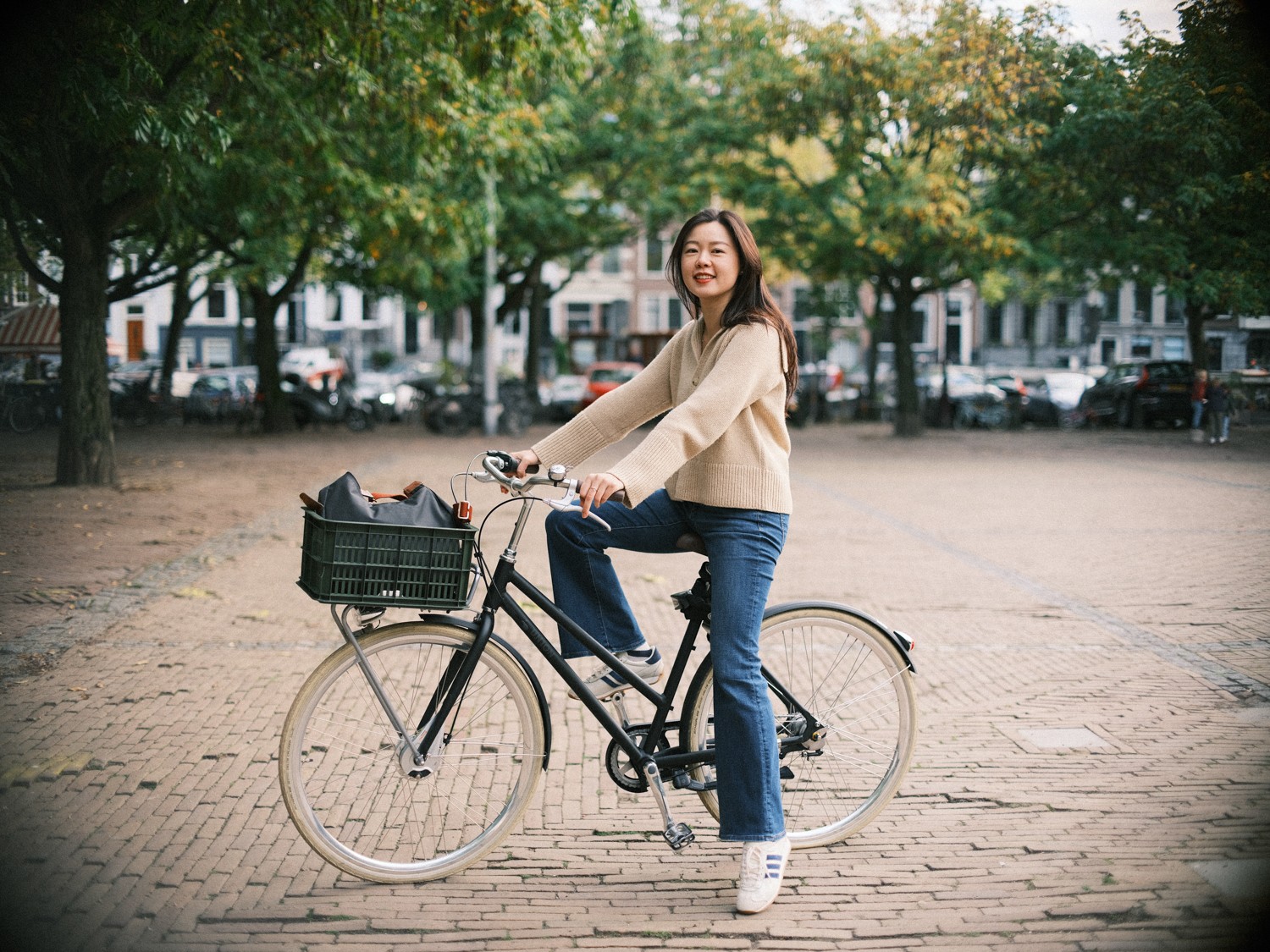 Vrouw op een zwarte fiets met één voet op de klinkers lacht naar de camera, met herfstbomen en geparkeerde auto’s erachter