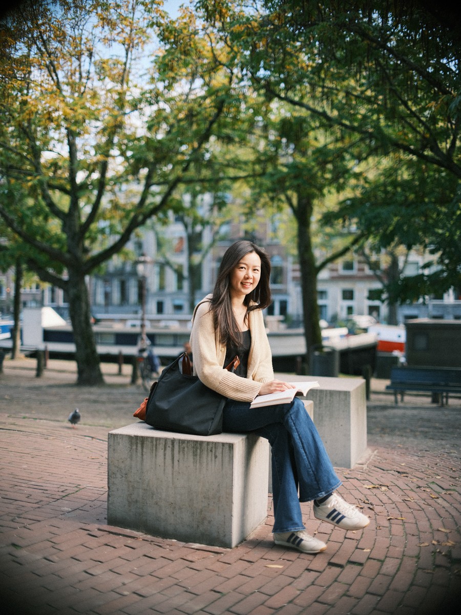 Vrouw lacht op een betonnen blok met een open boek op schoot, een duif op de klinkers en herfstbomen erachter