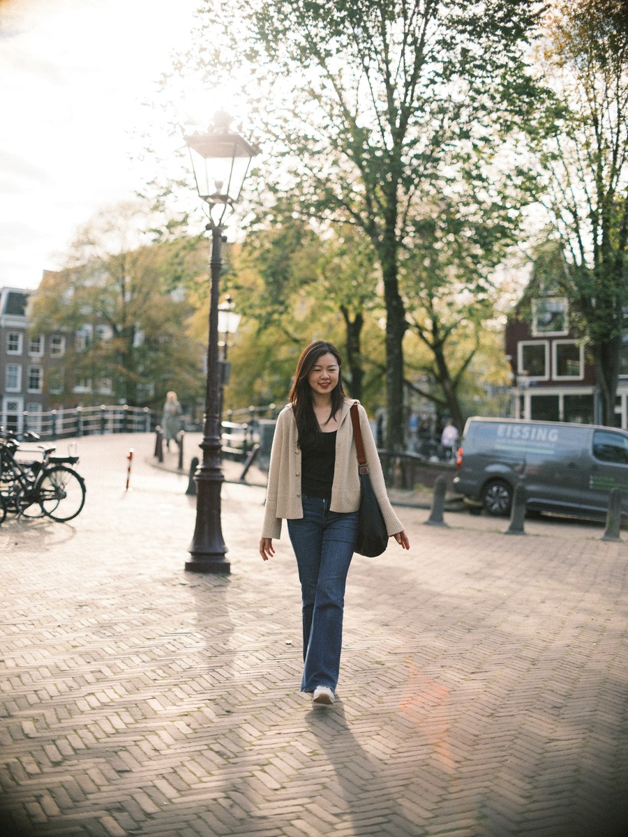 Vrouw in een beige vest en wijde jeans loopt naar de camera toe over klinkers in visgraatverband, met lage zon door de bomen