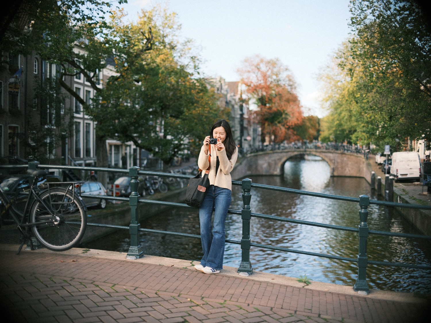 Soloreiziger leunt op een groen hek langs de gracht om een foto te maken met haar compacte camera, met een boogbrug en herfstbomen