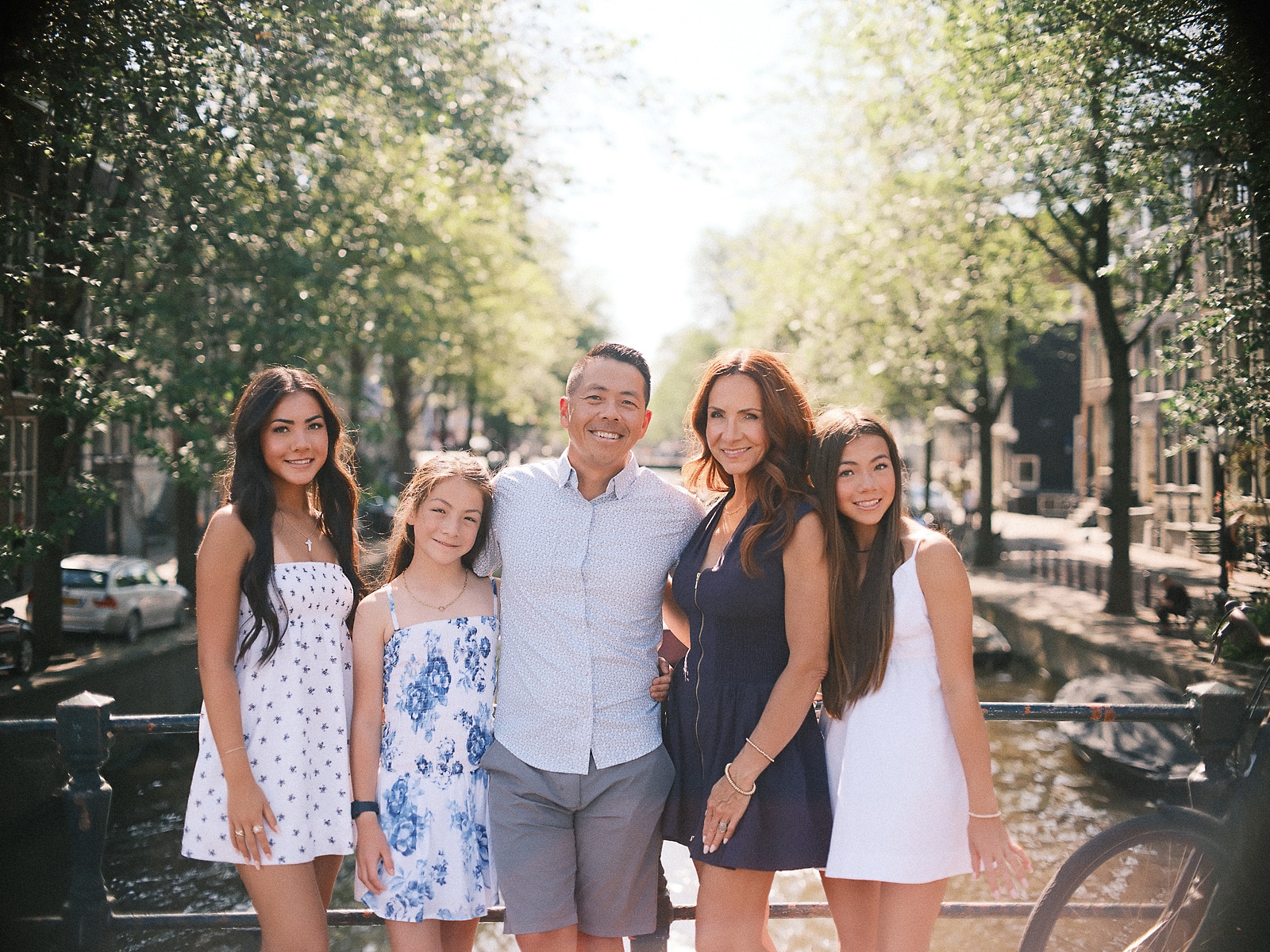 Gezin van vijf staat arm in arm op een grachtenbrug onder groene zomerbomen, een natuurlijke familiefotoshoot in Amsterdam