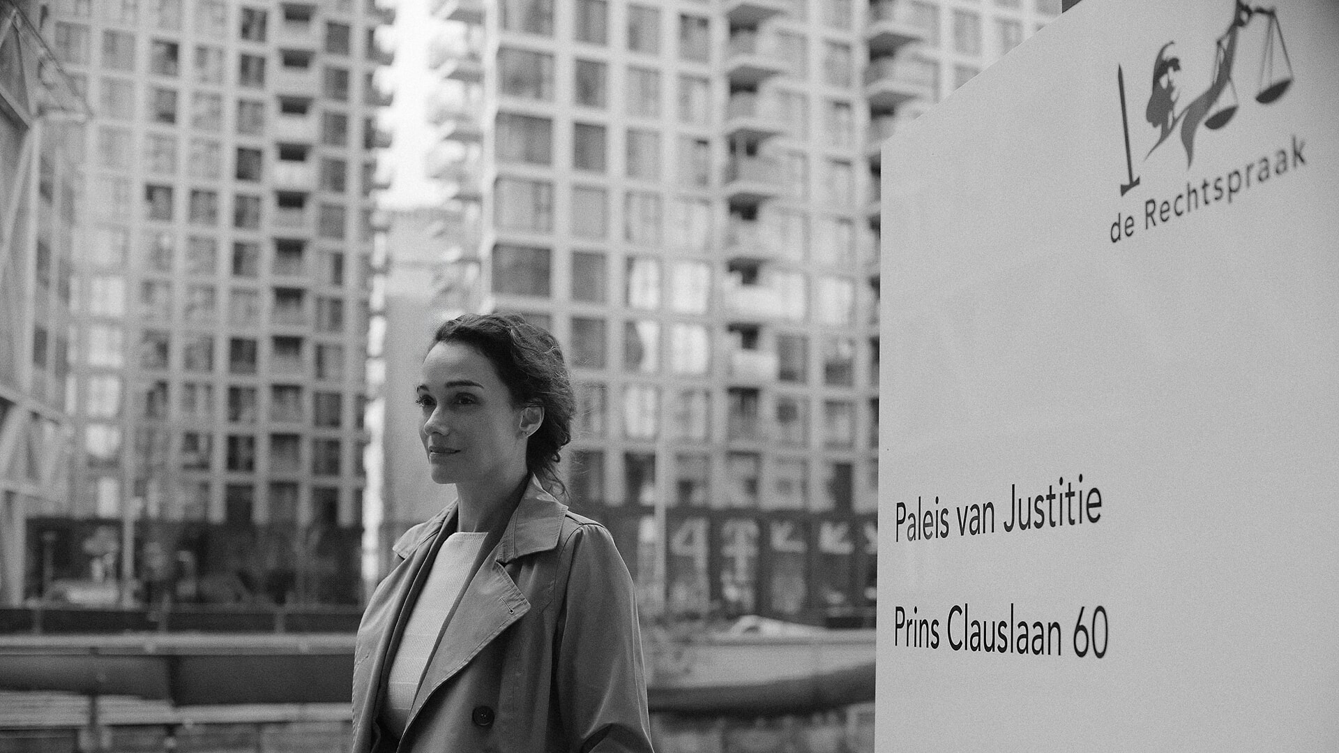 Vrouw in trenchcoat voor het Paleis van Justitie naast het naambord, met een hoge moderne gevel achter haar, een zwart-wit zakelijk portret