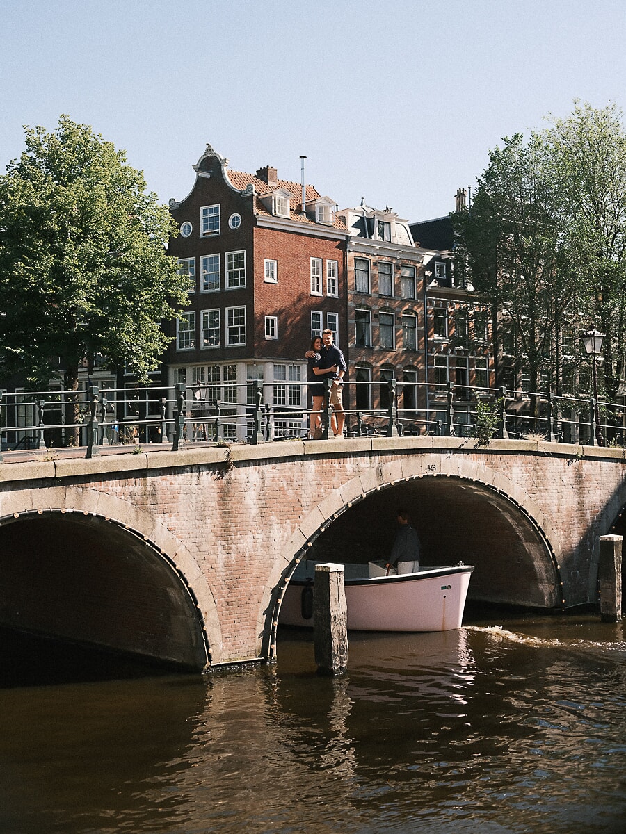 Couple embracing on a stone canal bridge as a boat passes beneath during their Amsterdam loveshoot
