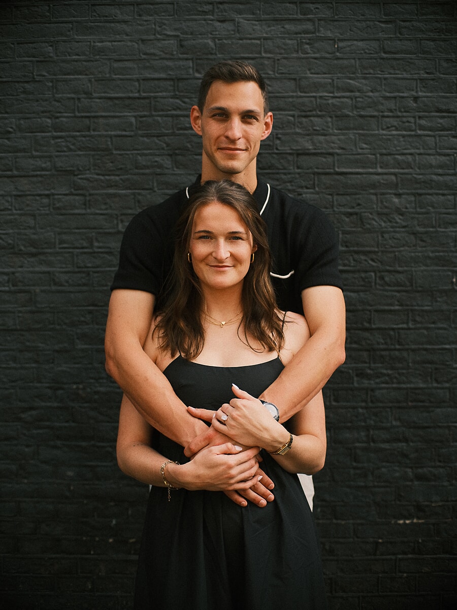 Engaged couple against a black brick wall, his arms wrapped around her with her engagement ring showing