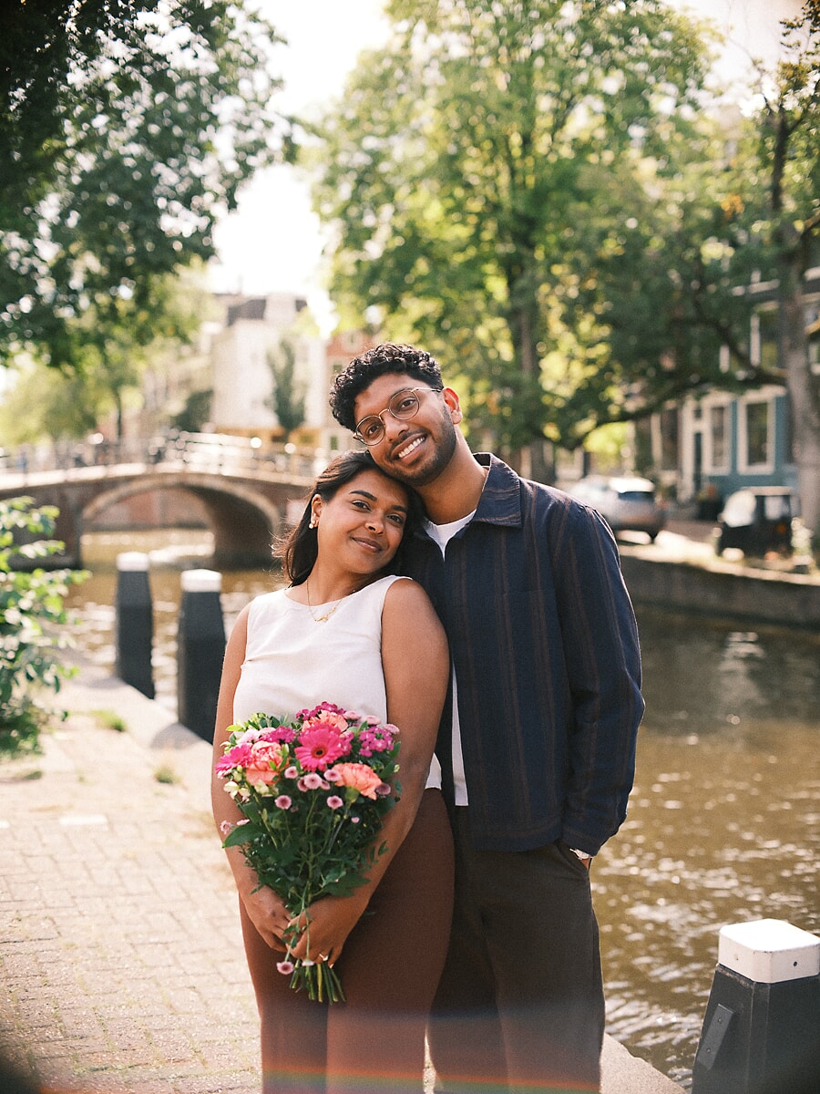 Engaged couple standing close beside an Amsterdam canal with a bouquet of pink gerberas, an arched bridge behind them