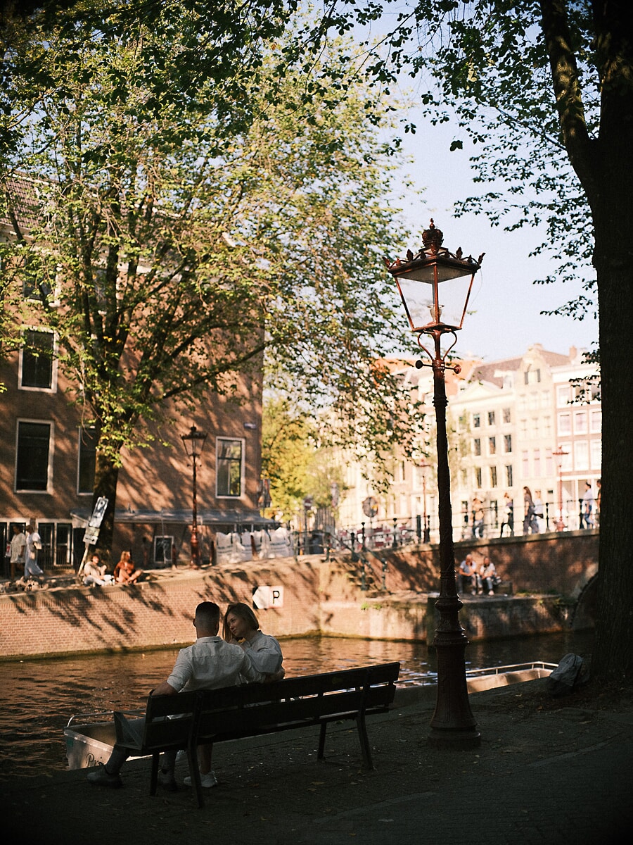 Engaged couple sitting close on a canal-side bench beneath an Amsterdam street lamp in the late afternoon