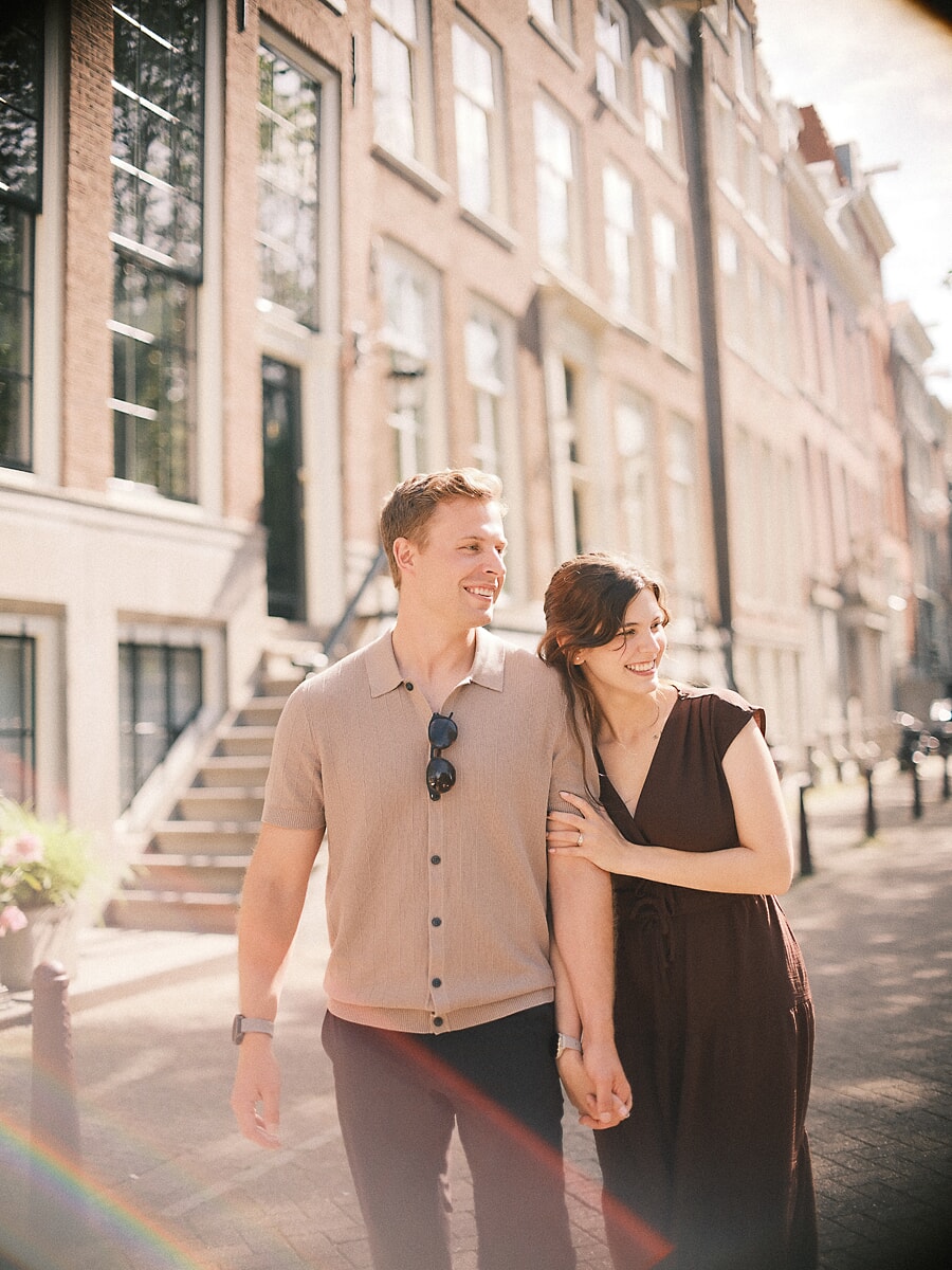 Engaged couple walking arm in arm along a canal street, the gentle glare of an artistic lens across the frame