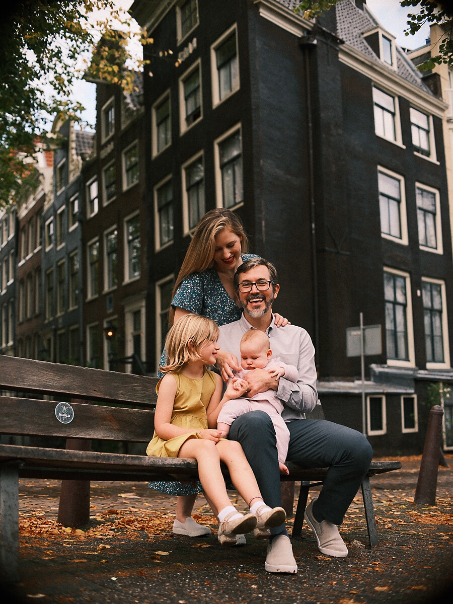 Family of four laughing on a bench in front of dark Amsterdam canal houses, a warm autumn family portrait