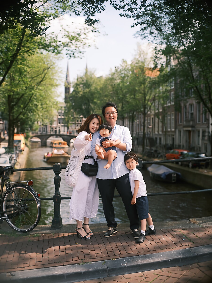 Family of four on an Amsterdam canal bridge in summer, a tour boat passing under the arch behind them