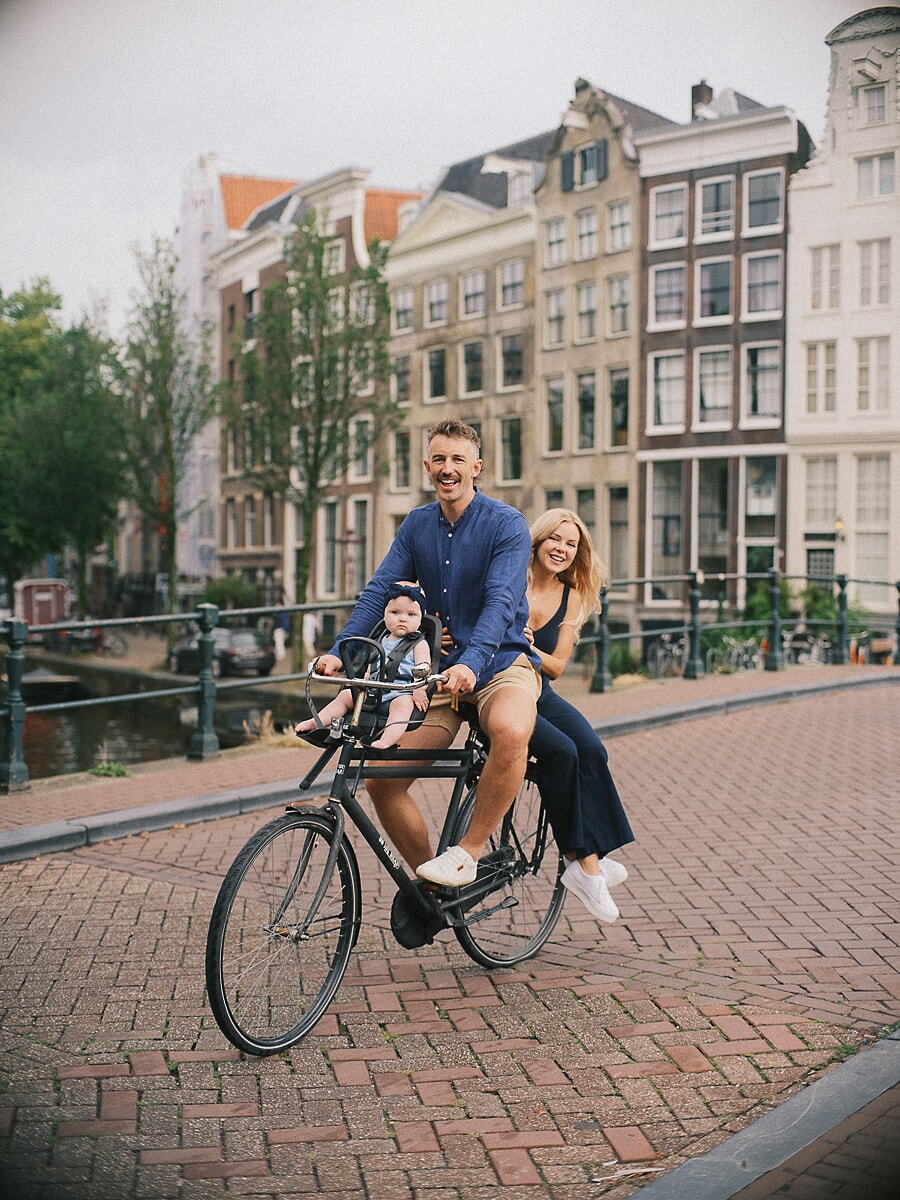 Parents cycling along an Amsterdam canal with their baby in the bike seat, a real-life moment from a family photo session
