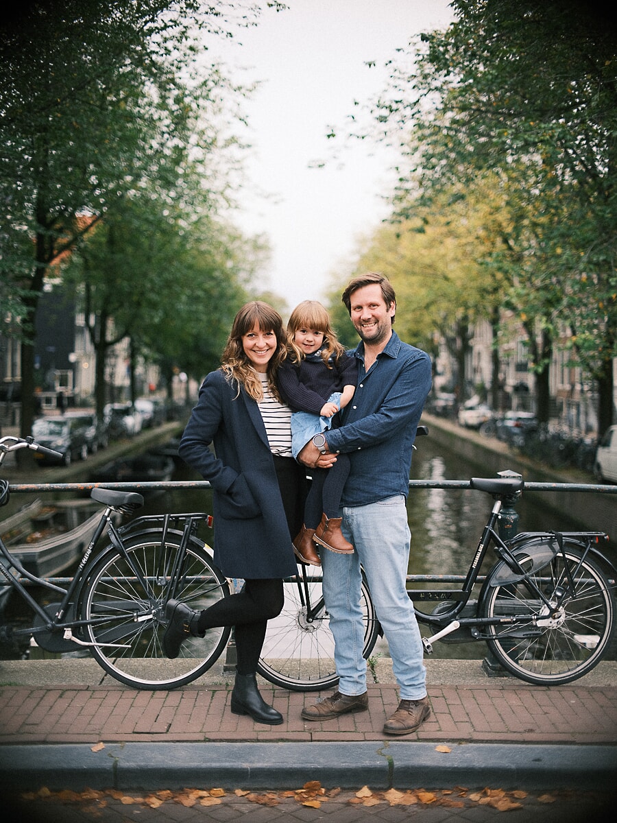Family of three on an Amsterdam canal bridge between parked bicycles, their daughter held between them