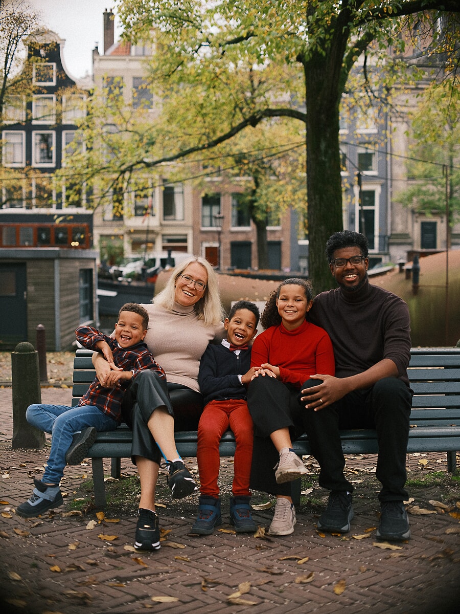 Family of five squeezed onto a canal-side bench in autumn, a family portrait in Amsterdam with a houseboat behind