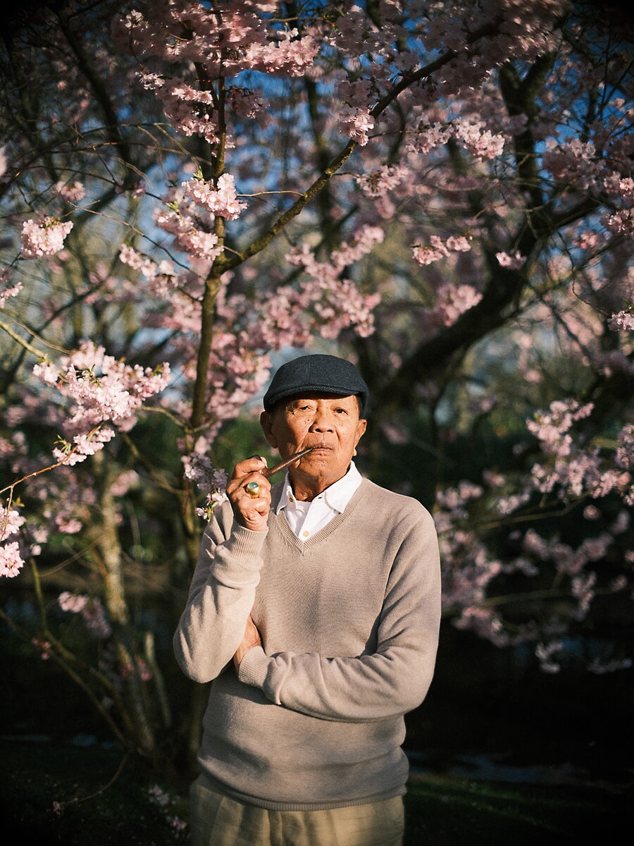Grandfather in a flat cap drawing on his pipe beneath pink cherry blossom, from a family anniversary in Amsterdam
