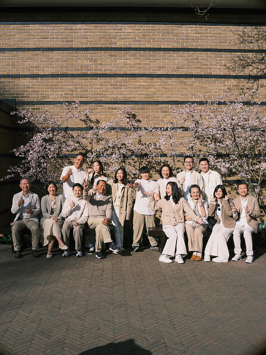 Fifteen family members in matching cream outfits under cherry blossom in Amsterdam, a group portrait at their anniversary shoot