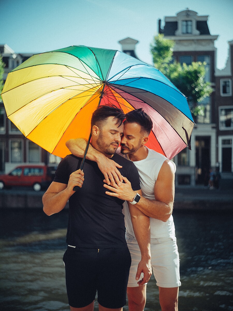 Two men embracing under a rainbow umbrella beside an Amsterdam canal, a vibrant partner portrait
