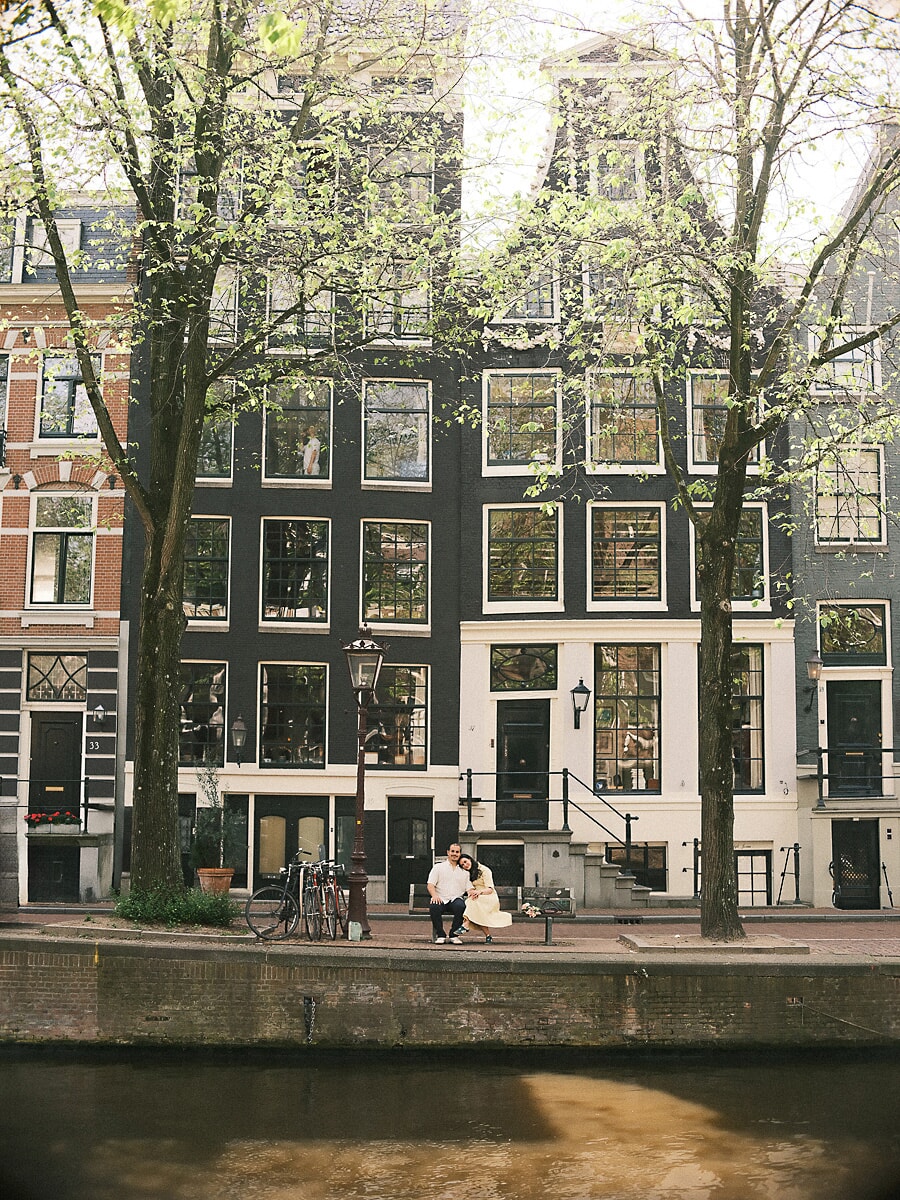 Couple on a bench beneath spring trees in front of Amsterdam canal houses, a wide frame from their loveshoot