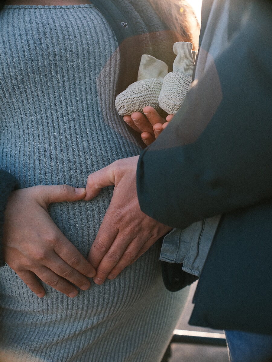Close-up of hands framing a bump in a grey knit dress while holding a pair of knitted baby booties