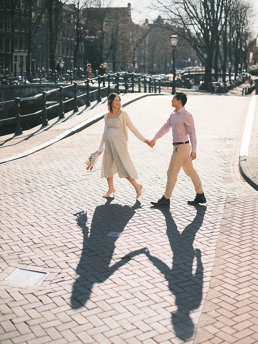 Expecting parents walking hand in hand across sunlit brick in Amsterdam, their long shadows stretching beside them