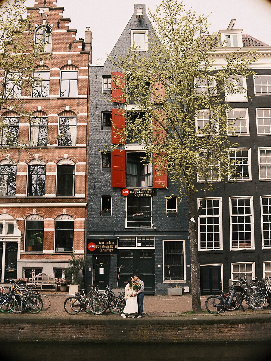 Expecting parents kissing among the bicycles below a tall Amsterdam canal house with red shutters, waiting on their first child