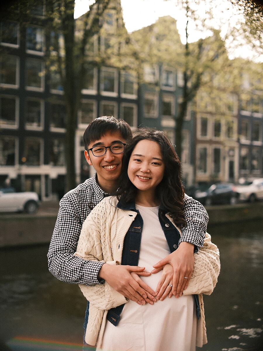 Expecting parents forming a heart with their hands on the bump as their family grows, Amsterdam canal houses behind them