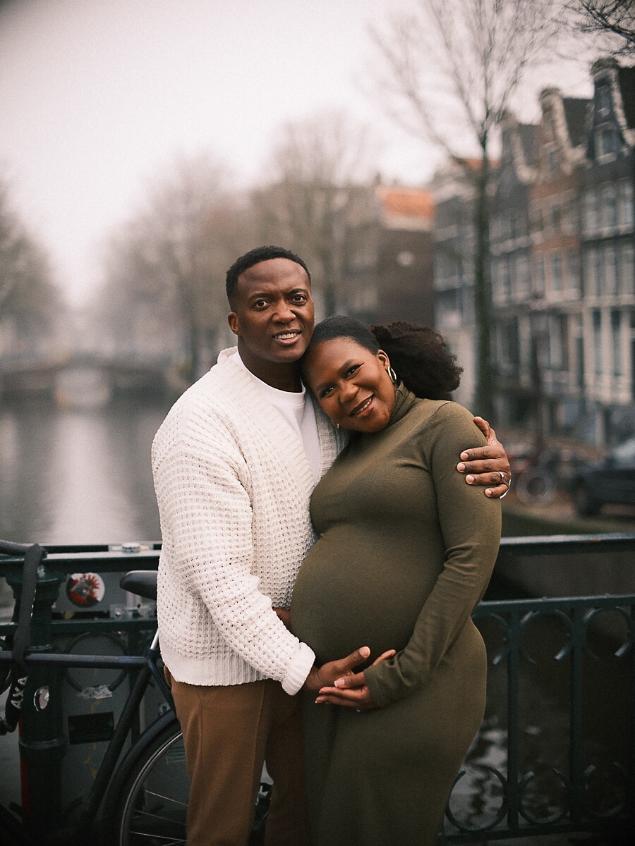 Expecting parents holding the bump on a misty Amsterdam canal bridge, quiet maternity photography in winter