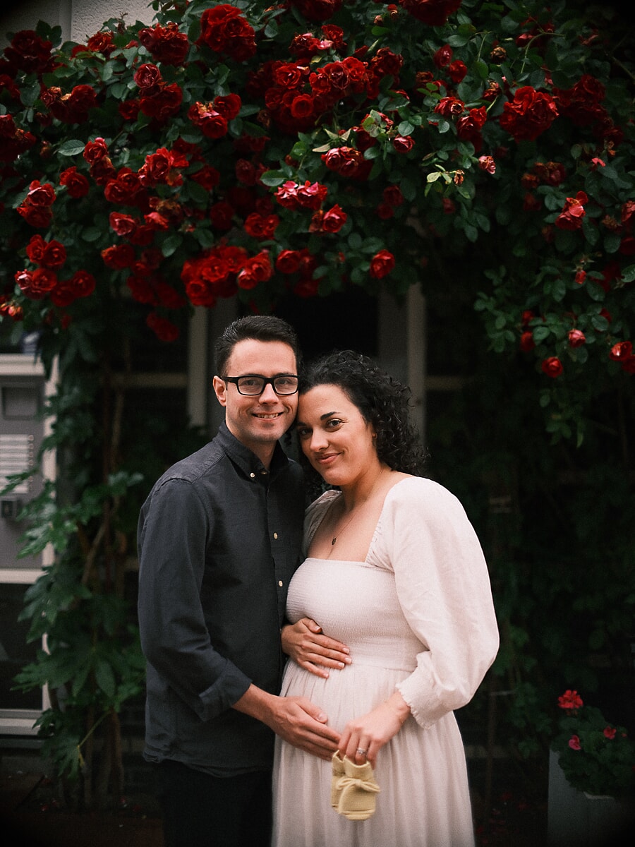 Expecting couple beneath a wall of red climbing roses, a pair of baby shoes in her hand