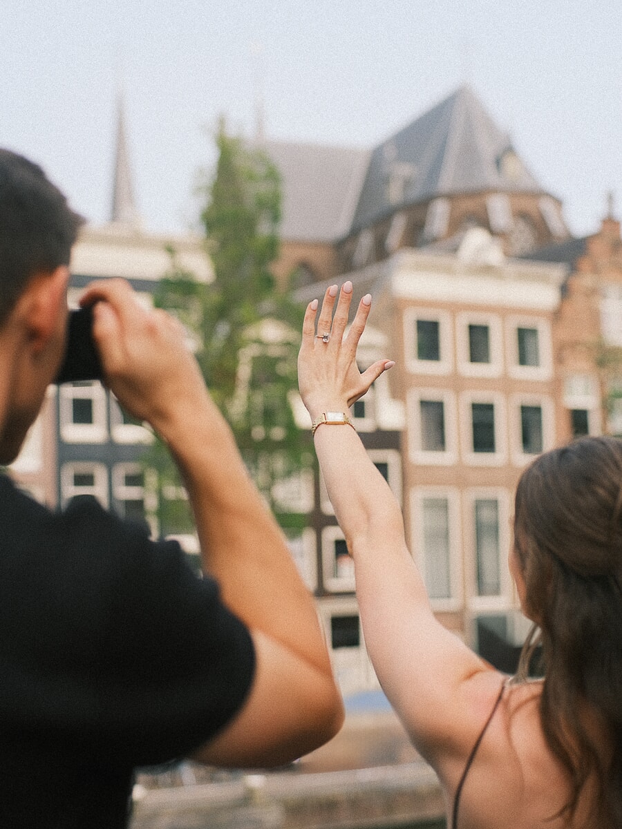 Newly engaged woman raising her hand to show the ring while her partner photographs her by Amsterdam canal houses