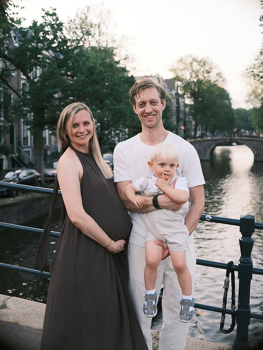 Family of three by an Amsterdam canal at dusk expecting their fourth, an arched bridge behind them