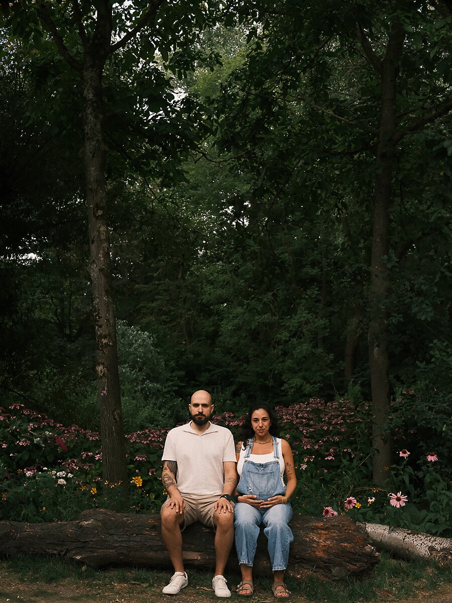 Expecting couple seated on a fallen log among summer flowerbeds during a pregnancy photoshoot in an Amsterdam park