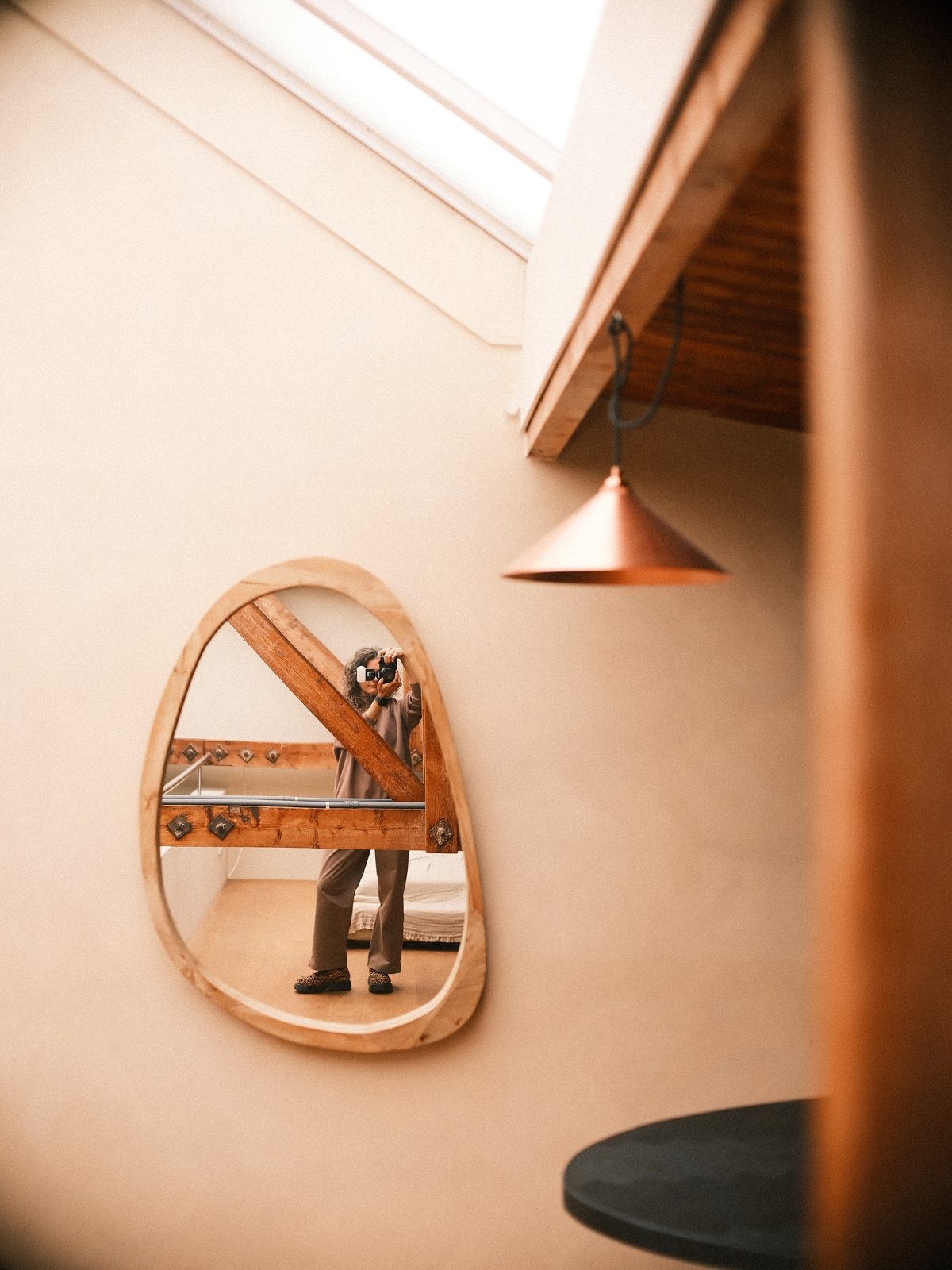 Aleksandria Rudenko reflected in an egg-shaped wooden mirror, holding a camera to her eye in a warm attic under a skylight and a copper lamp