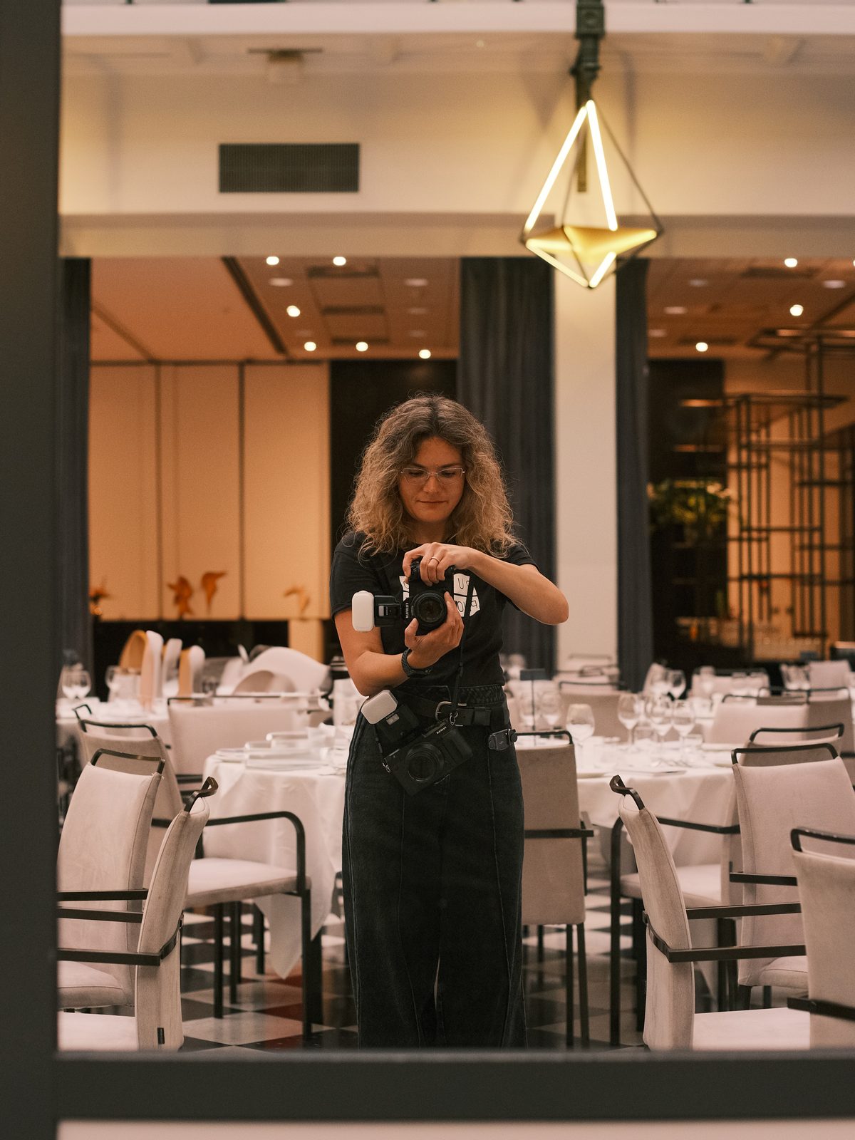 Aleksandria Rudenko reflected in a glass door, adjusting her camera in a ballroom set with white tables and chairs under a golden pendant light