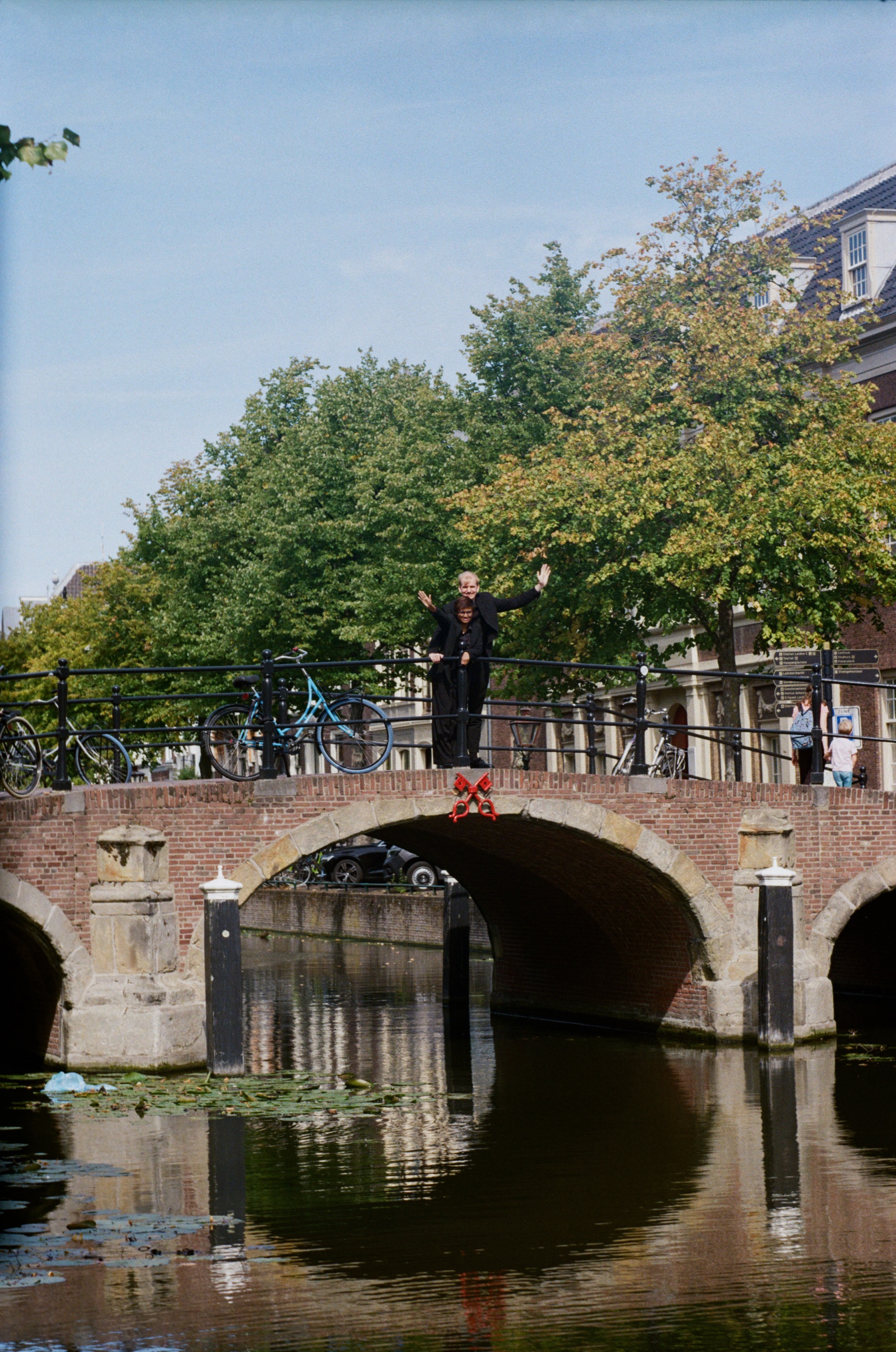 Arched brick bridge mirrored in still canal water, a man in black waving from the railing under a clear September sky, on color film