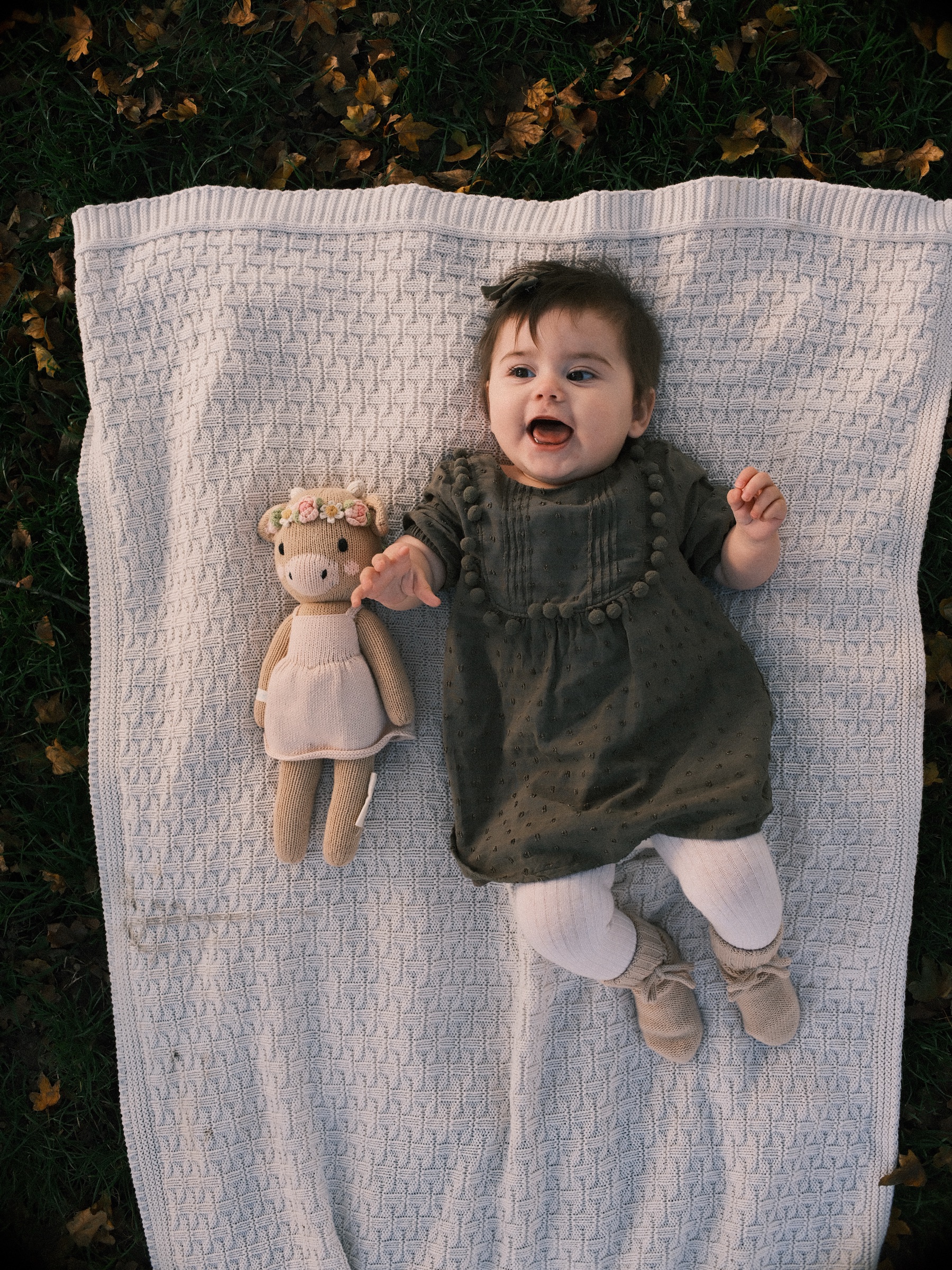 Seen from above, a smiling baby lies on a white knitted blanket next to her doll, her olive dress and white tights on the wool
