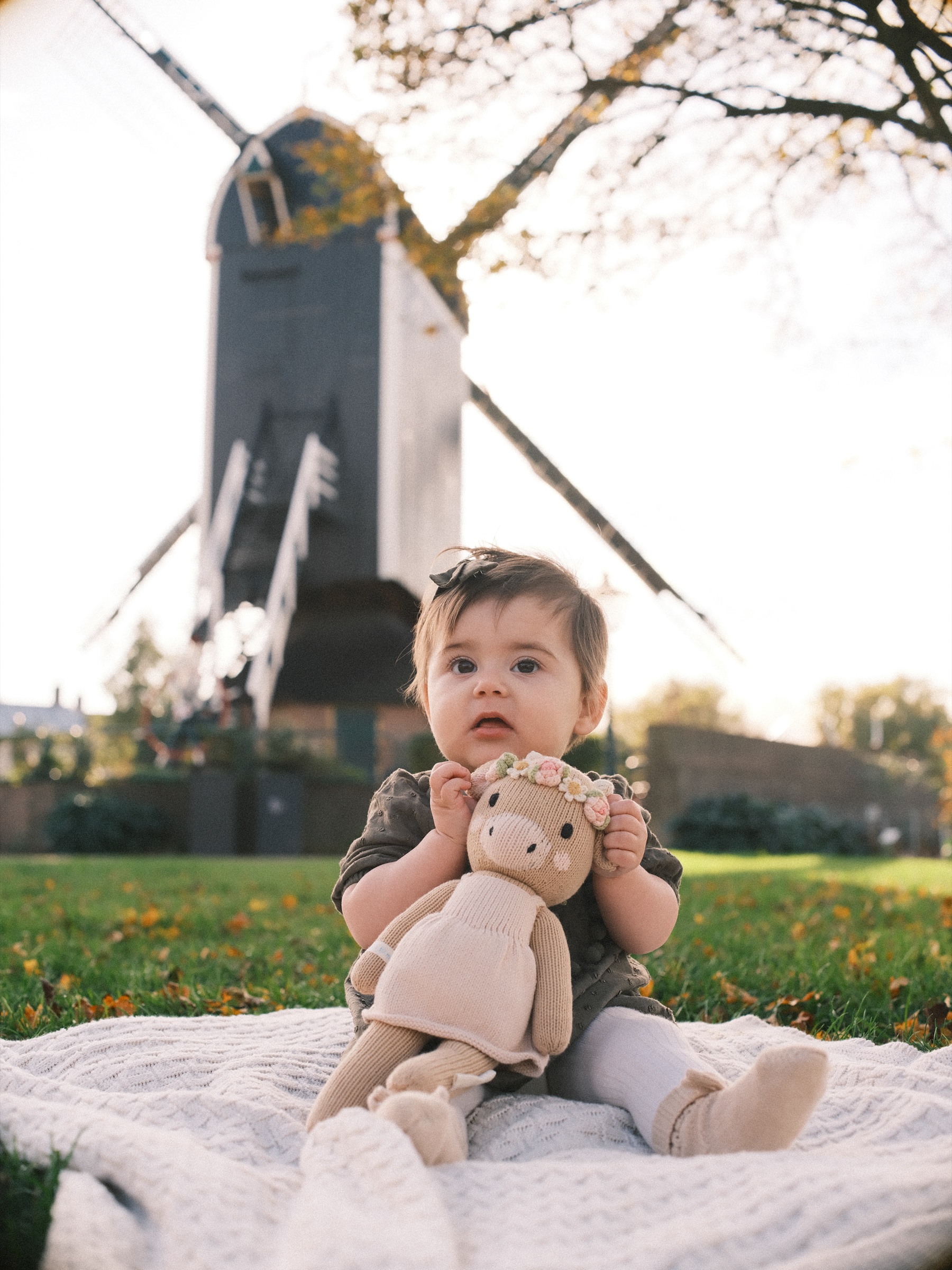 Baby girl holding a knitted doll in a cream dress on a white blanket, the dark wooden body of the windmill behind her