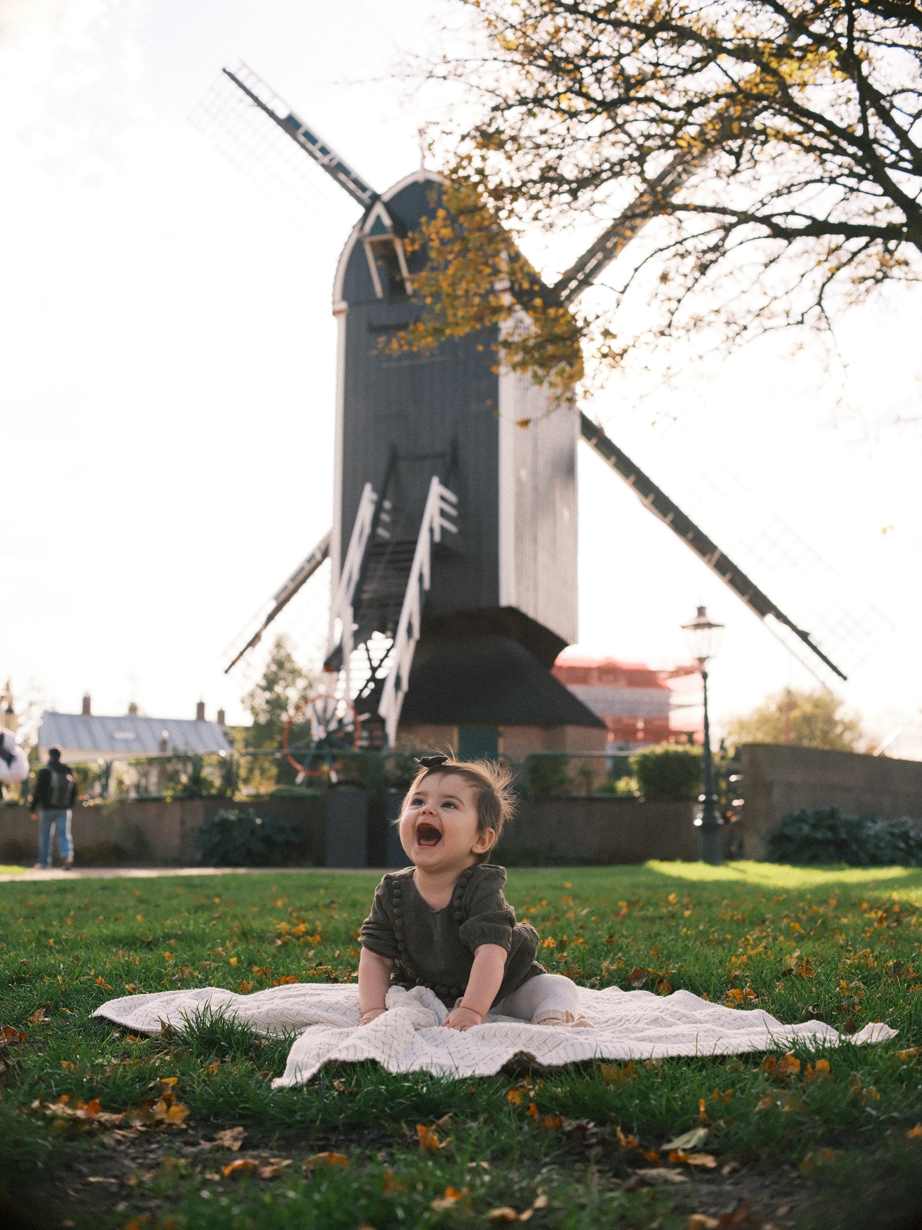 Baby girl in an olive dress laughing on a white blanket in the grass, the wooden post mill De Put rising behind her