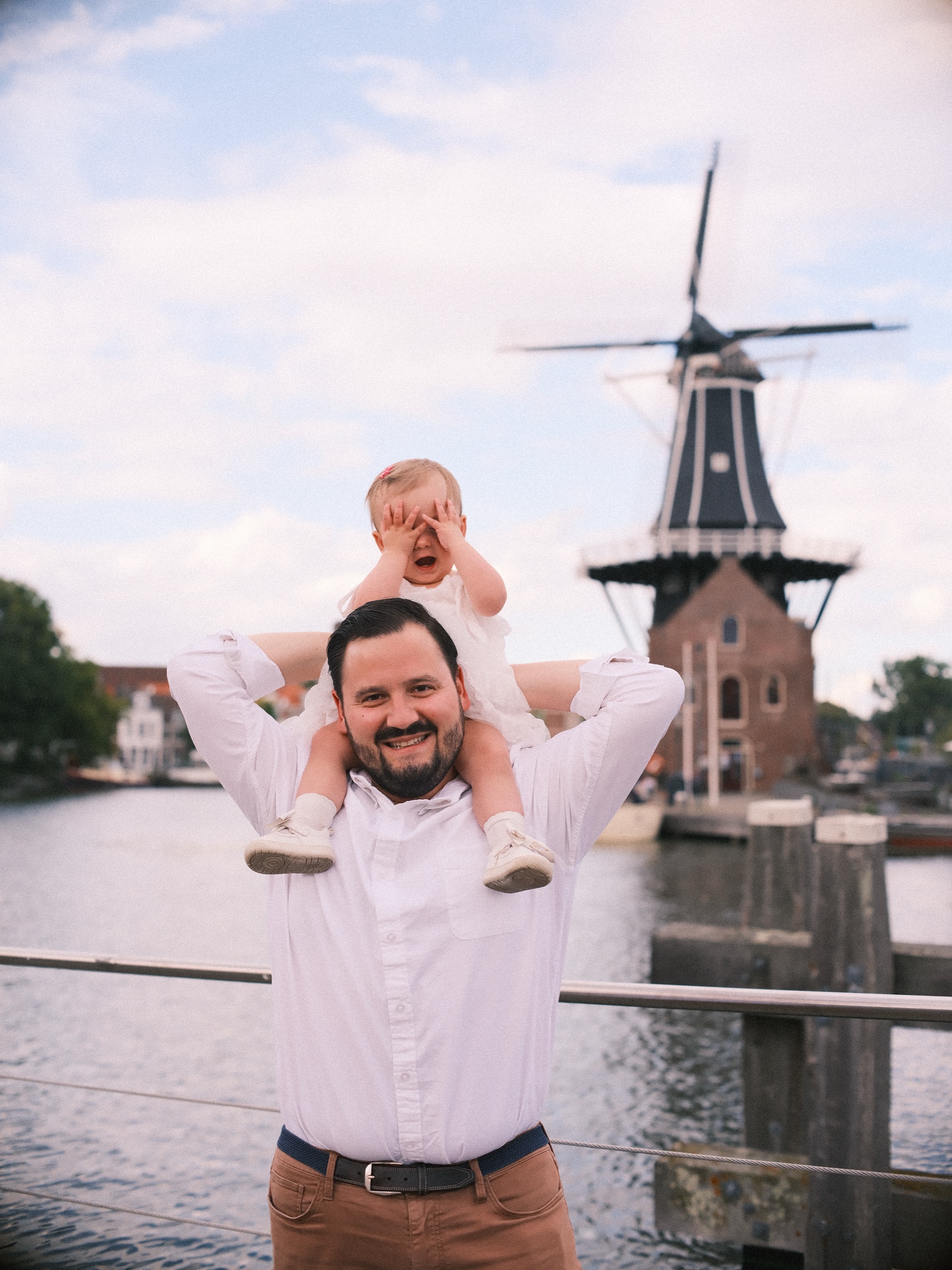 Lachende vader met zijn dochtertje op zijn schouders, dat haar handjes voor haar ogen houdt, met molen De Adriaan erachter