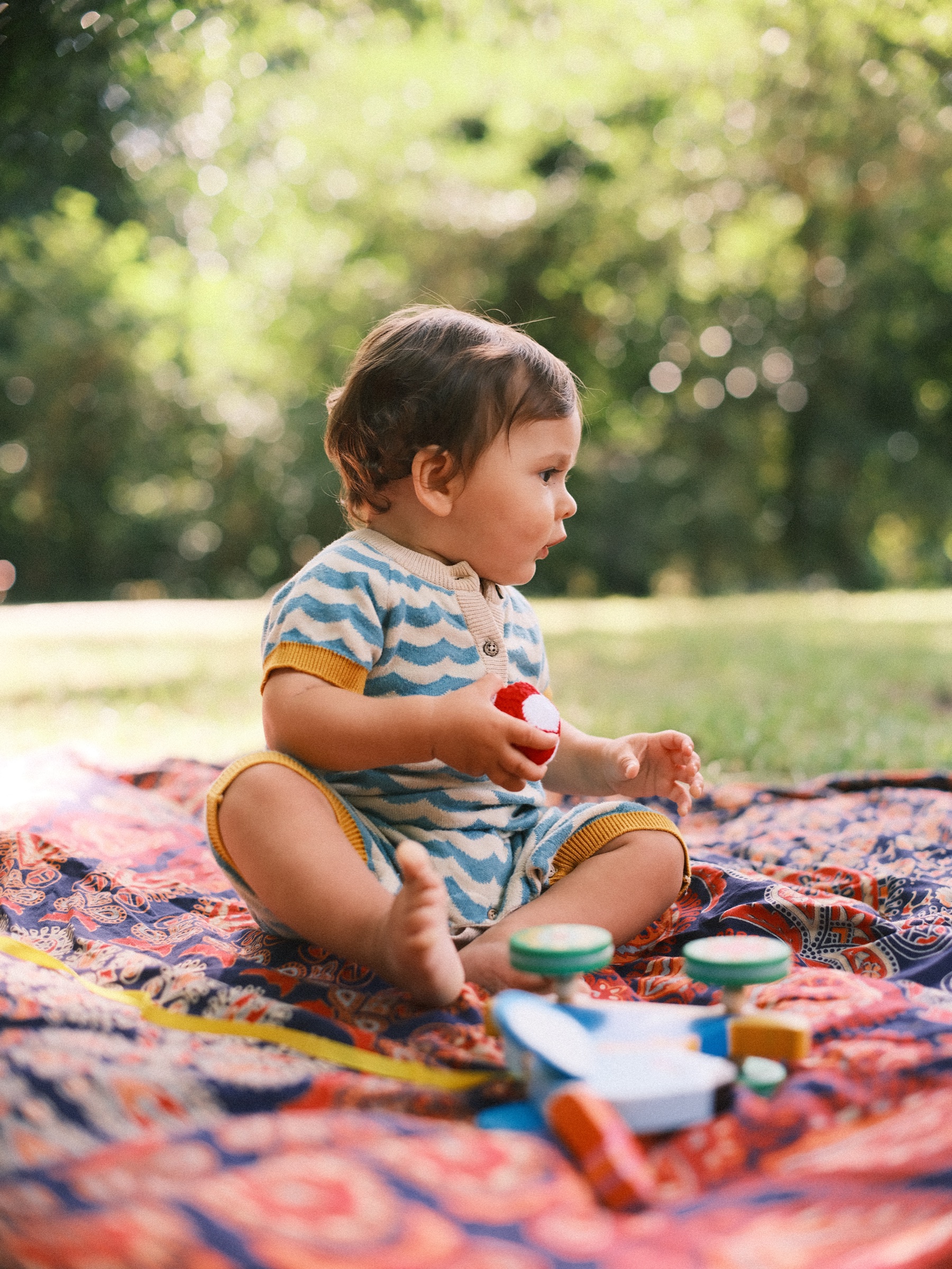 Baby zit op het kleed van de picknick tussen houten speelgoed, met een klein rood-wit balletje in zijn handen