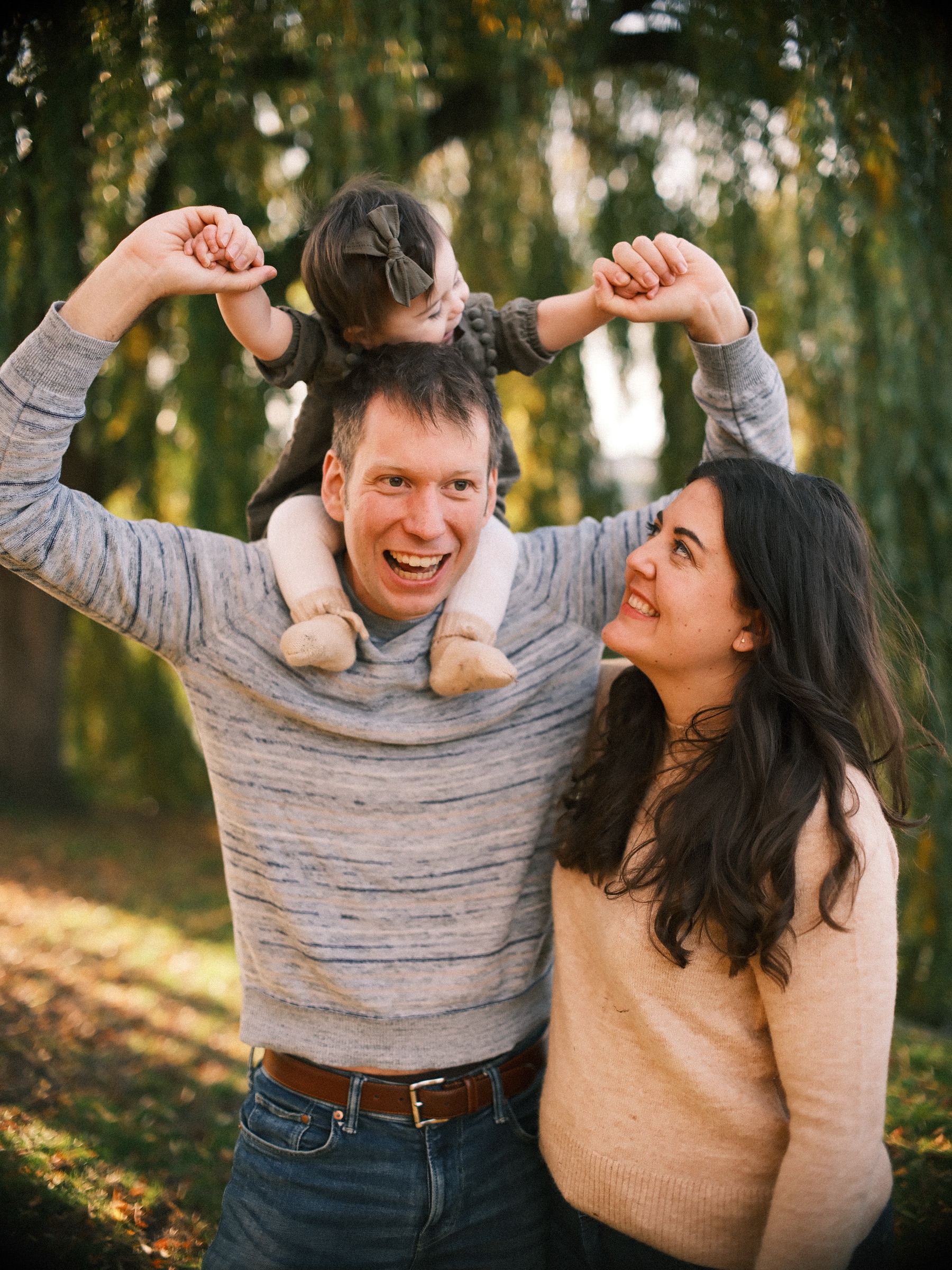 Baby on her father's shoulders holding his hands up high while her mother looks up at her, both parents laughing by a willow