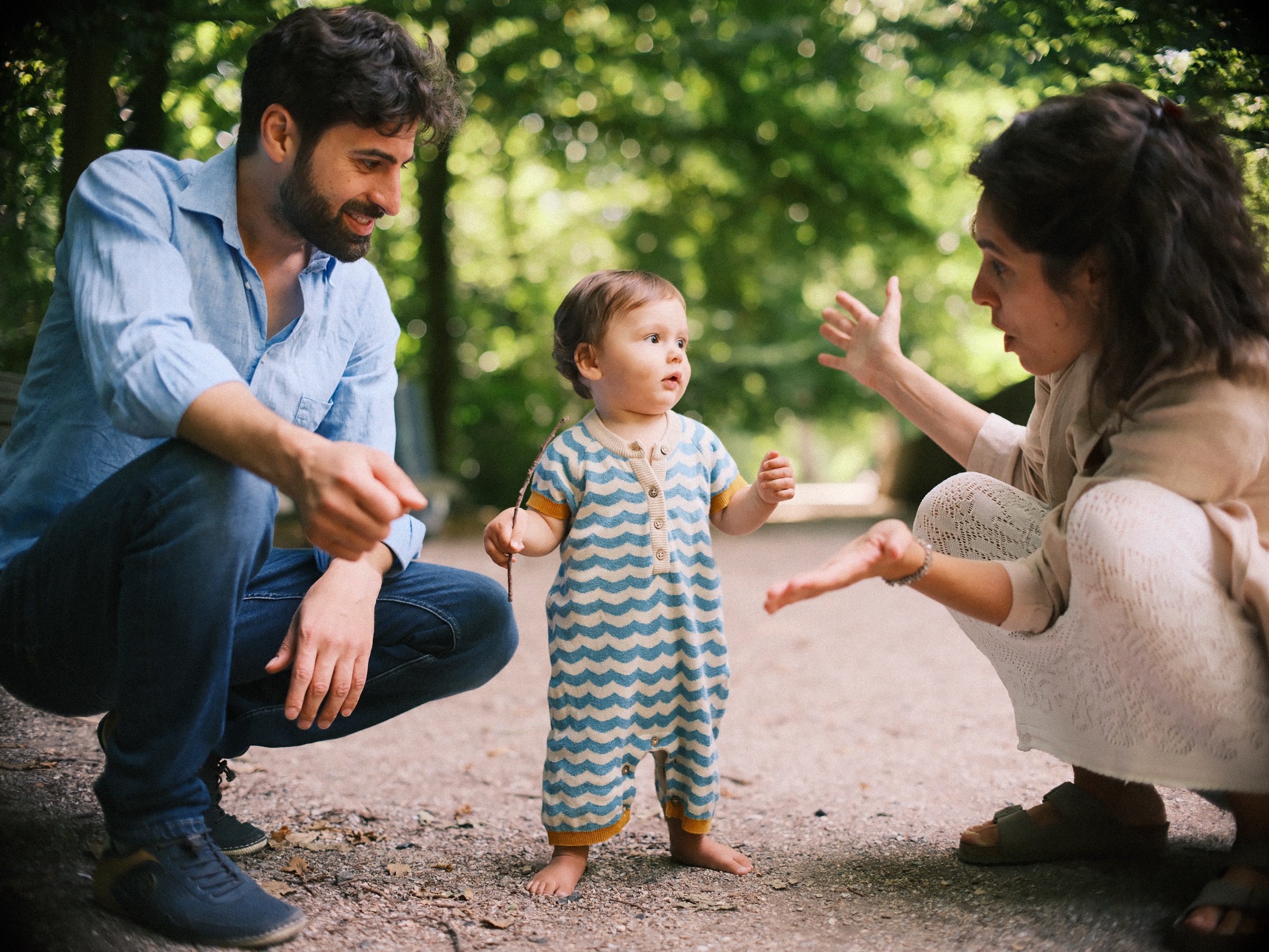 Baby in een gebreid pakje staat helemaal zelf op een grindpad, terwijl zijn ouders gehurkt hun handen klaarhouden