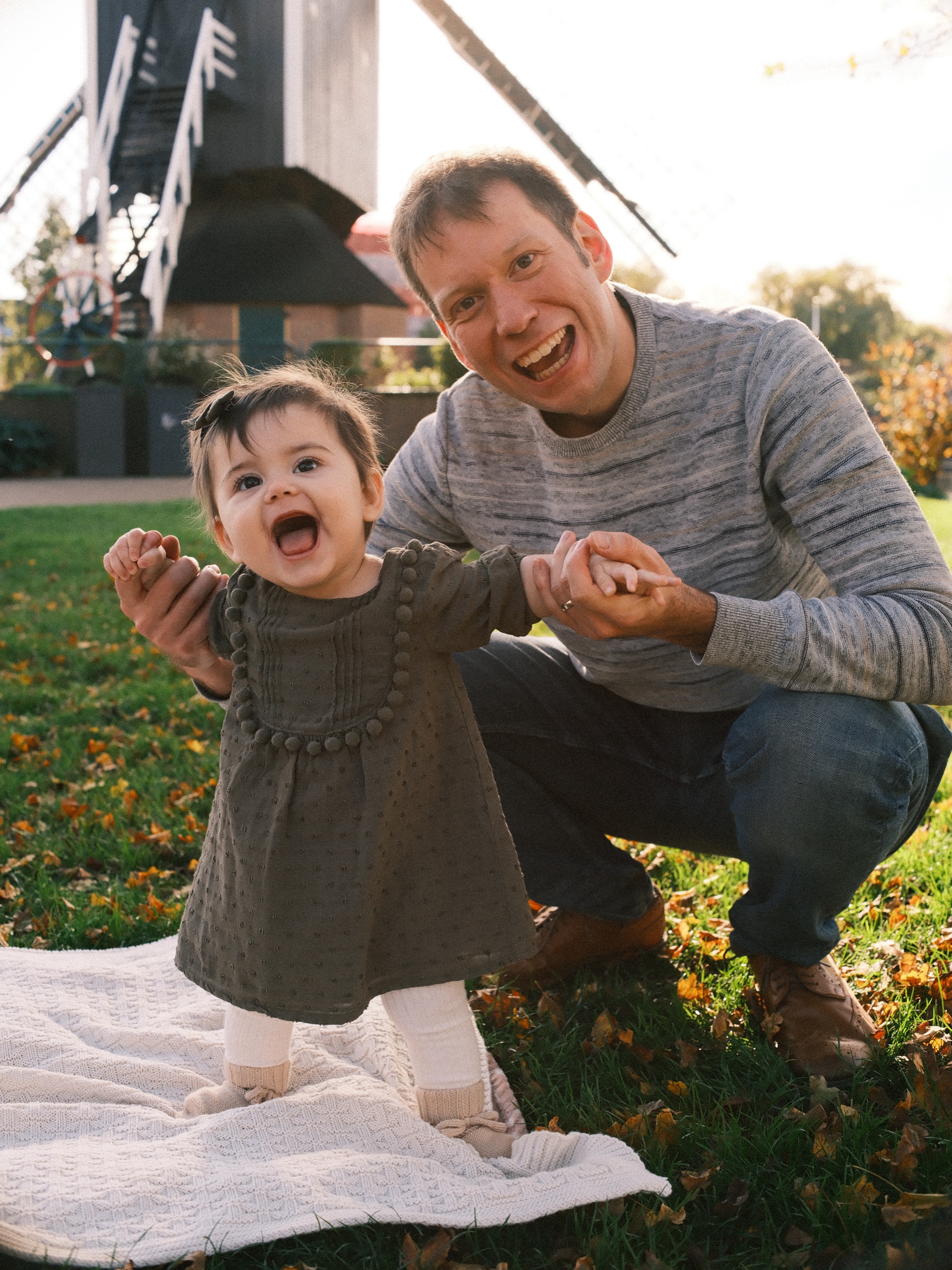 Baby girl standing with her father's help on the blanket, both laughing with open mouths below the mill in the autumn sun