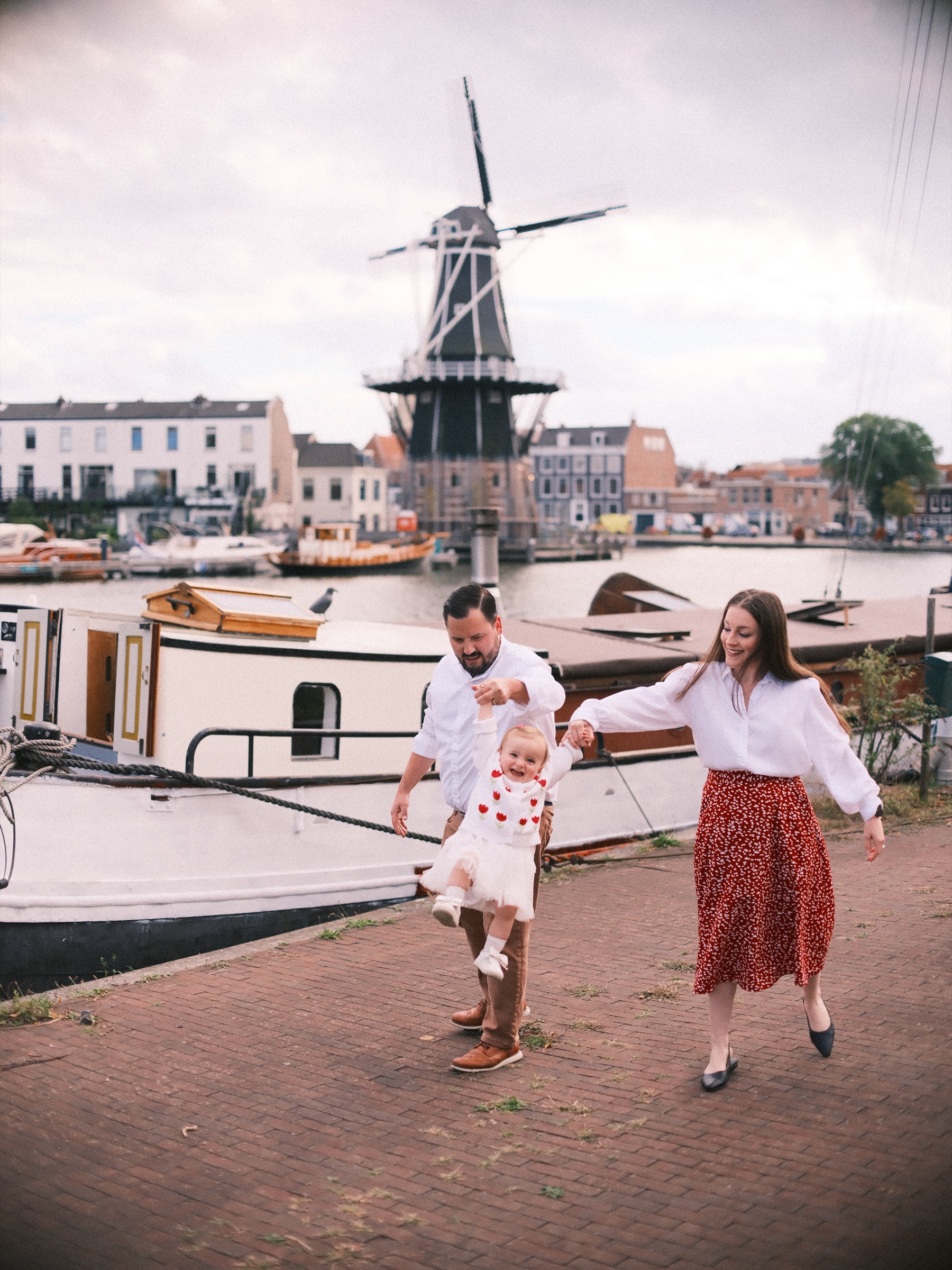 Ouders houden de handjes vast van hun dochter, die stapjes zet over een kade in Haarlem, met molen De Adriaan achter de boten