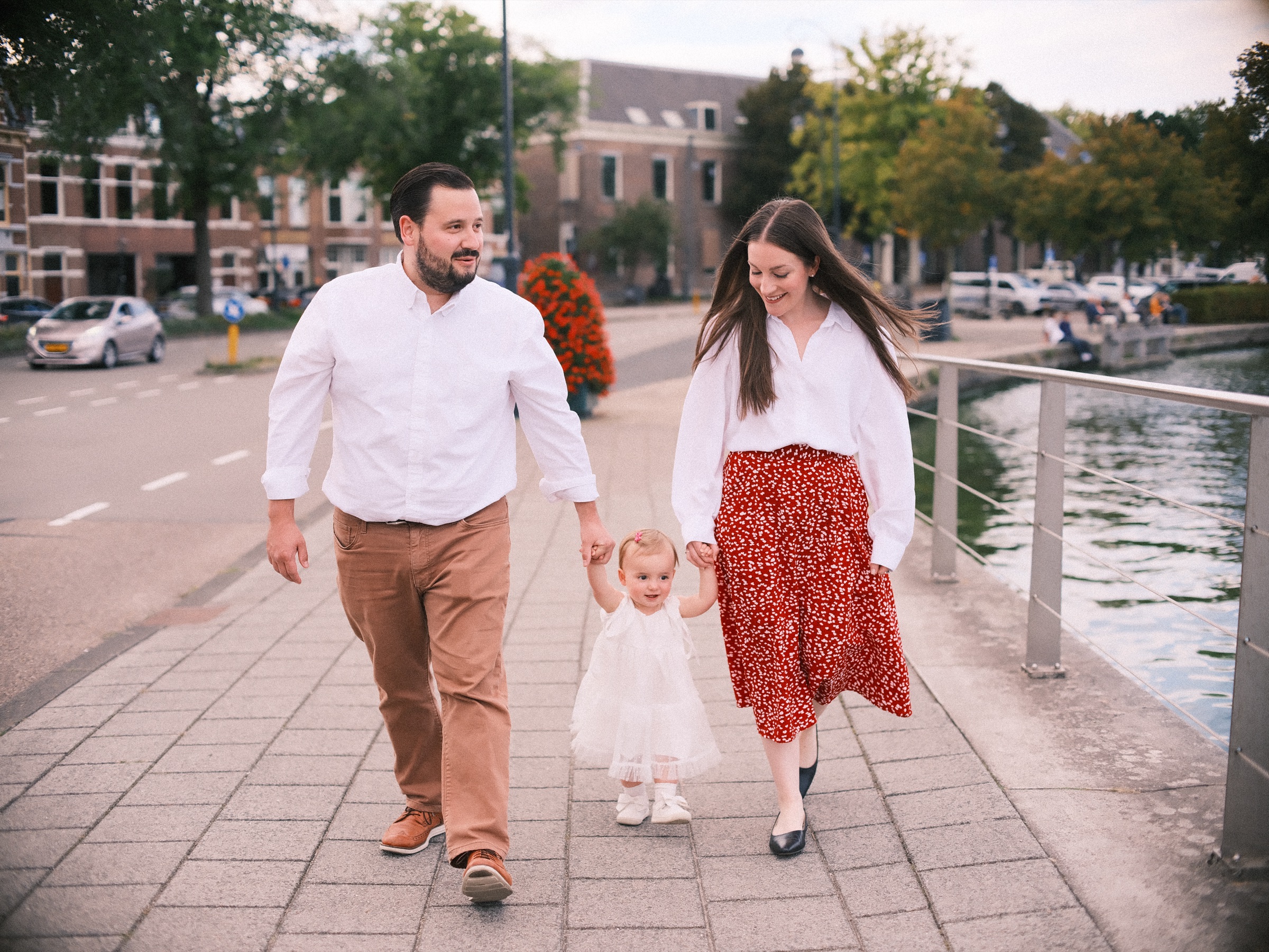 Dreumes in het wit zet haar eerste stapjes hand in hand met haar ouders over de wandelkade langs het Spaarne, bomen erachter