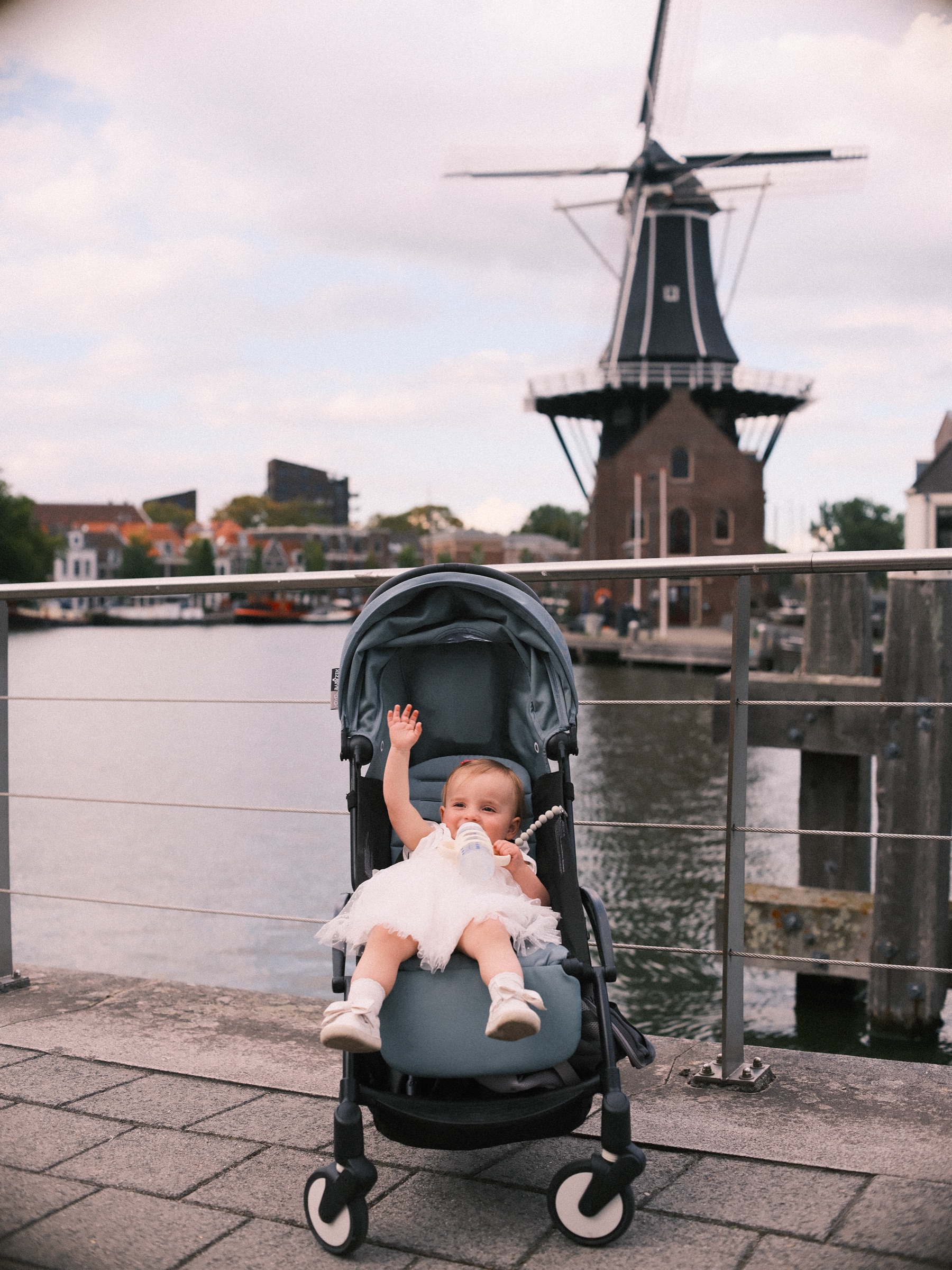 Dreumes in een grijze wandelwagen zwaait met één hand voor molen De Adriaan, met de reling en het water achter haar