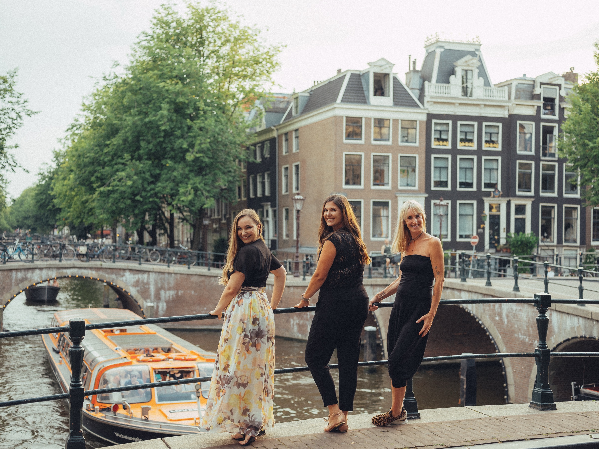 Bachelorette party of three leaning on a canal bridge railing as a tour boat passes below, tall Amsterdam houses behind
