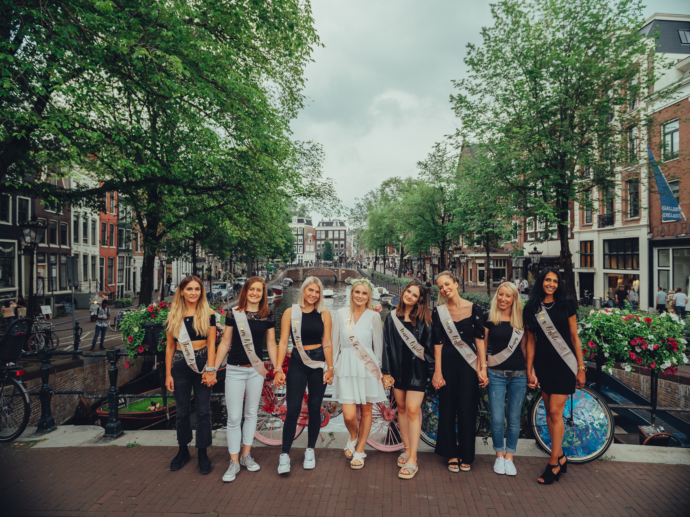 The bride and seven friends in sashes holding hands on a canal bridge in front of bikes covered in flowers, trees along the water